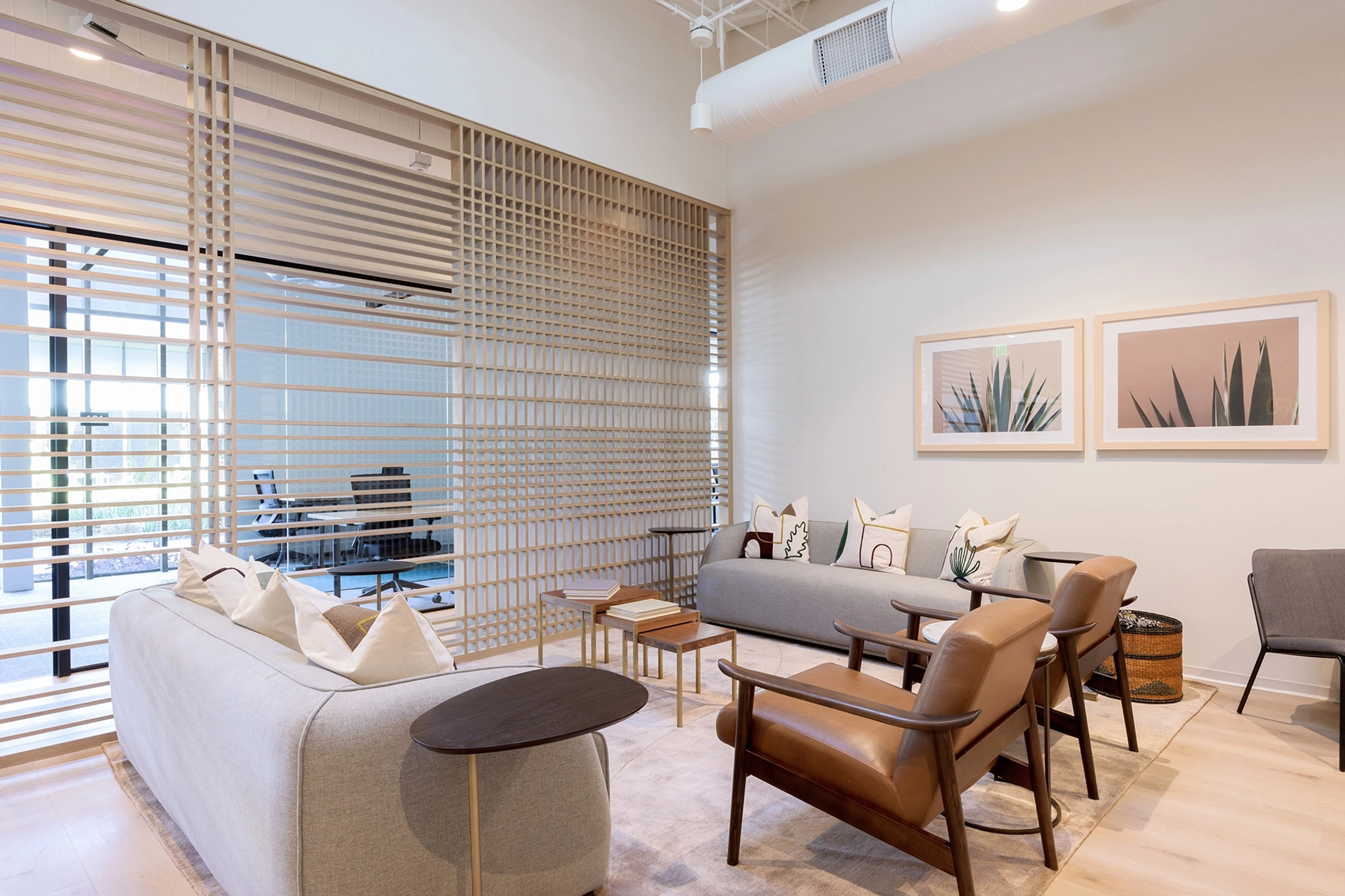 A modern office lounge area in Austin with two gray sofas, brown leather chairs, wooden tables, and wall art, separated from a meeting room by a wooden slatted partition.