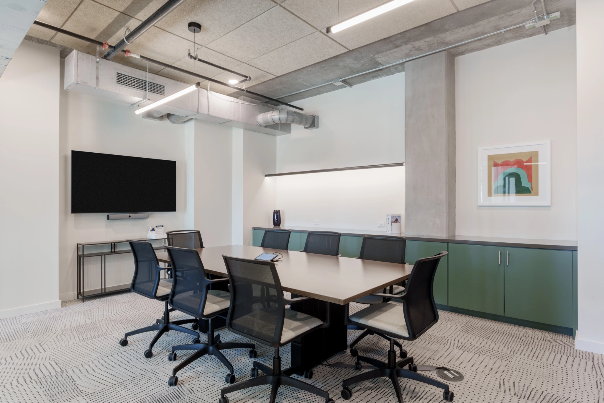 Modern conference room in an Austin office, featuring a rectangular table, eight black chairs, and a wall-mounted TV. Artwork adorns the walls beneath an industrial ceiling with exposed ducts, complemented by a patterned carpeted floor for a sophisticated workspace.