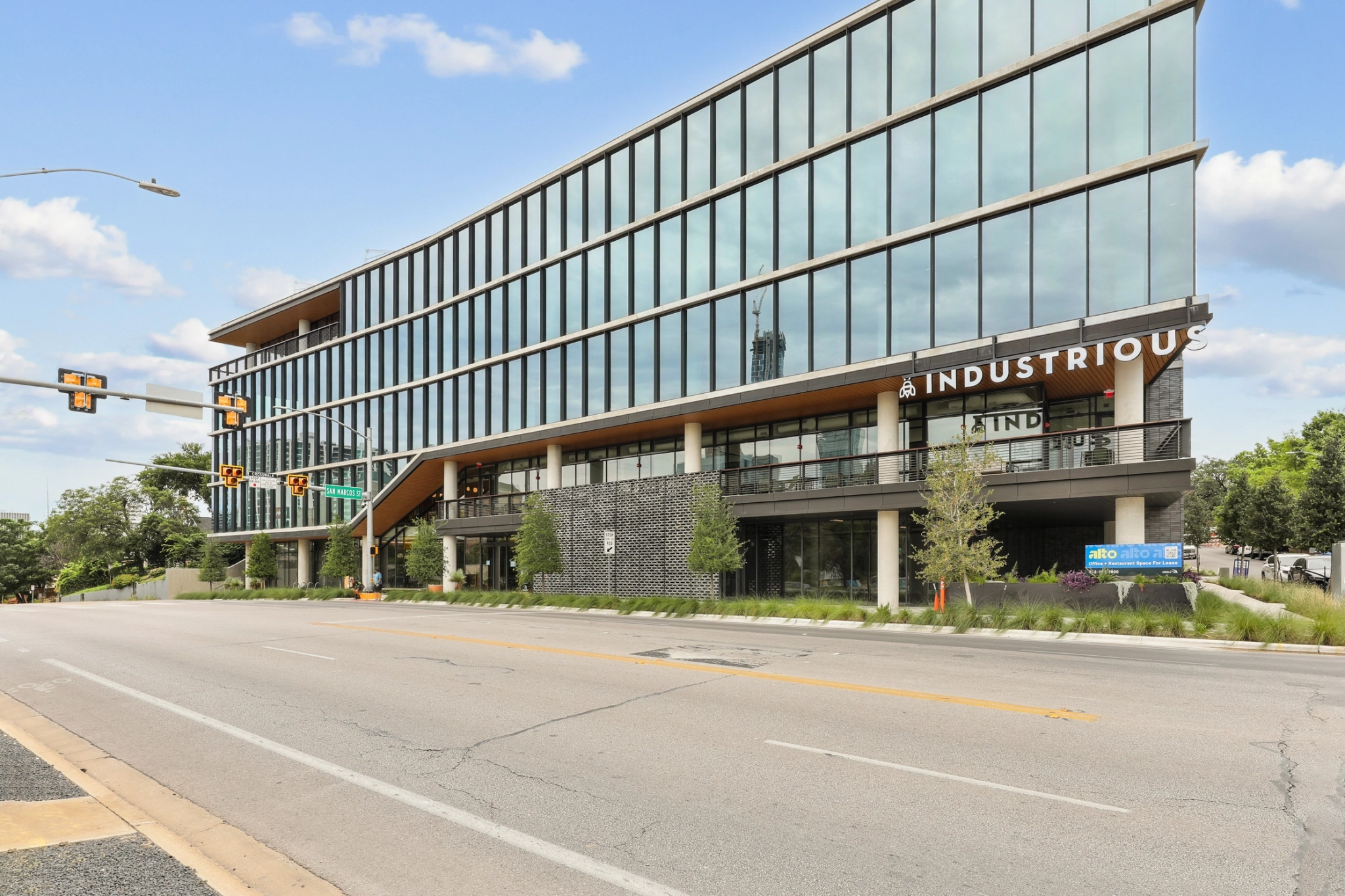 Un moderno edificio de oficinas con grandes ventanales y un cartel de "Industrious" ofrece espacios de coworking en la esquina de una calle con semáforos y zonas verdes cerca.
