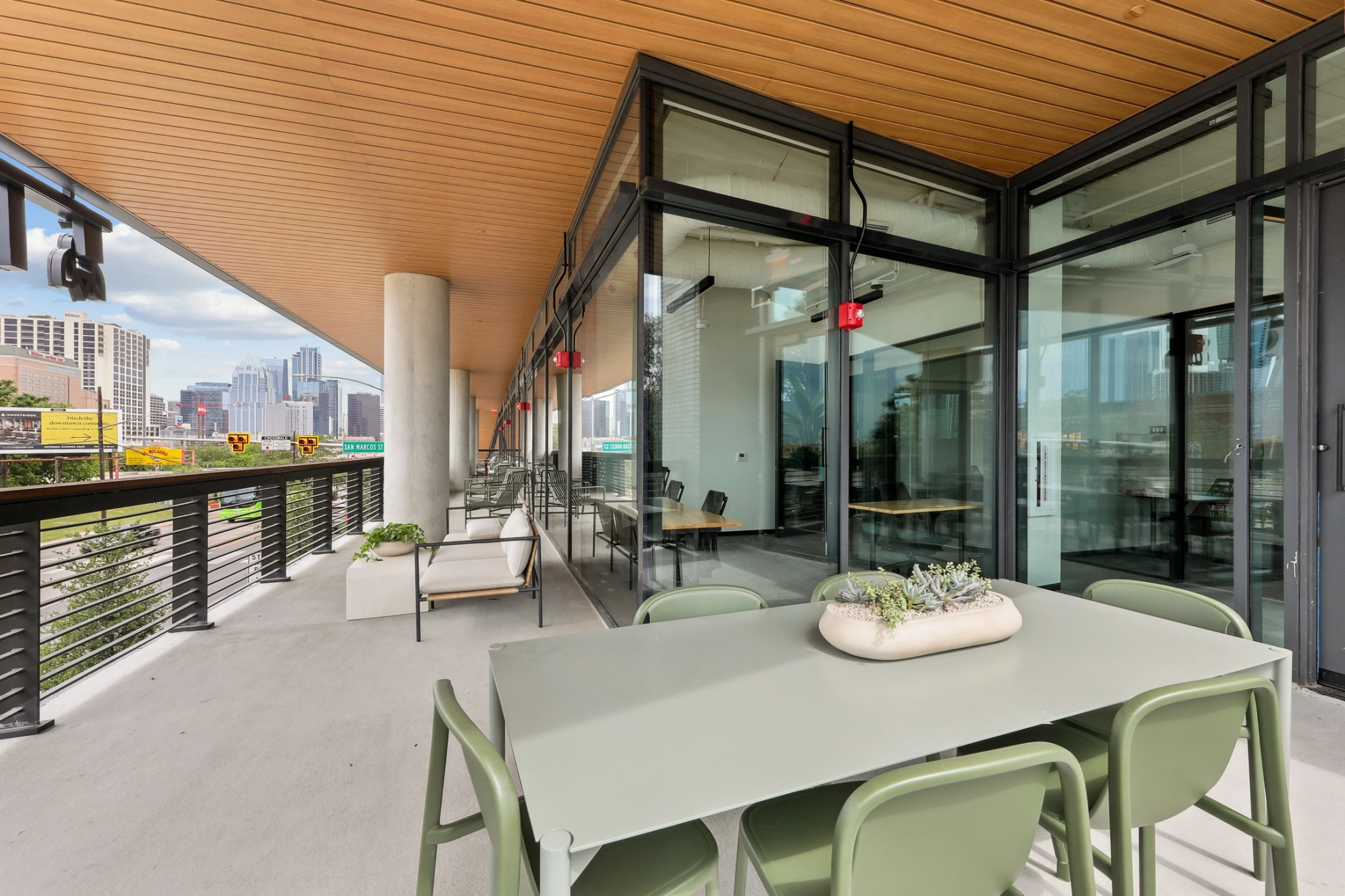 Modern outdoor balcony with a dining table, chairs, potted plants, and glass walls overlooking a cityscape—perfect as a stylish workspace or informal meeting room surrounded by greenery.