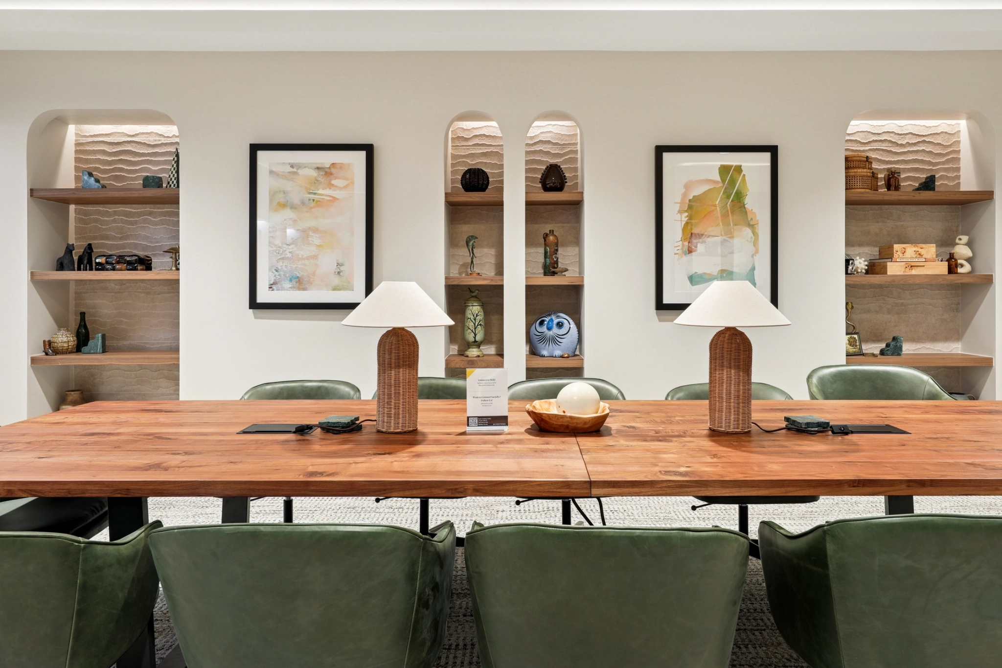 A modern Austin meeting room featuring a wooden table, green chairs, two lamps, wall shelves with decor items, and framed abstract art on a white wall—perfect for an inspiring workspace.