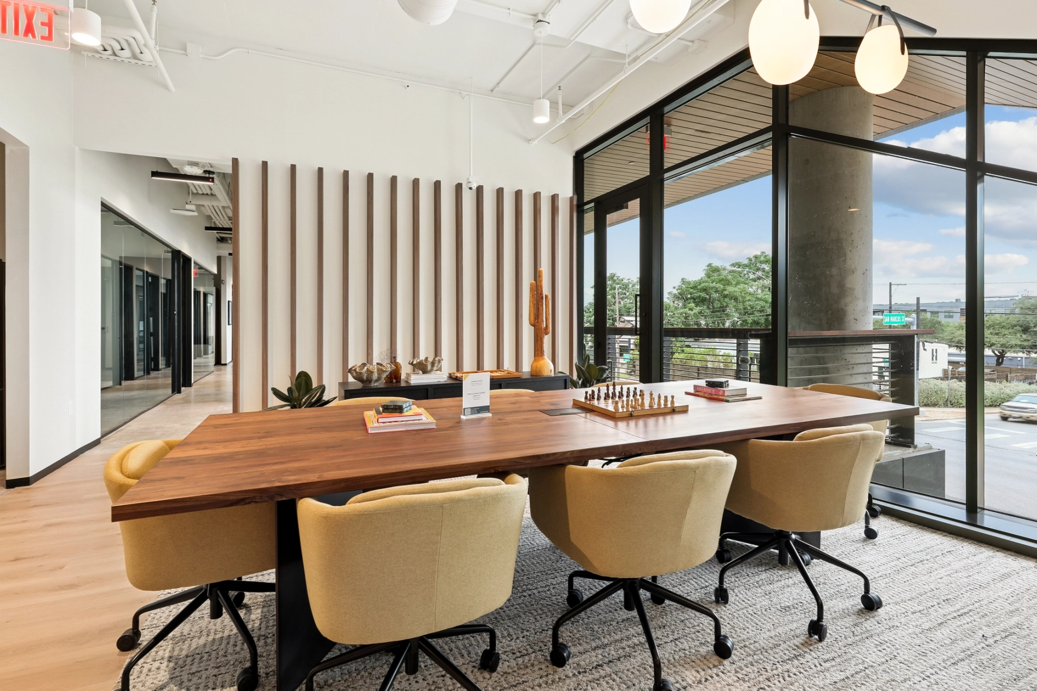 Un espacio de coworking moderno con una mesa de madera, seis sillas beige, muebles elegantes y grandes ventanales con vistas a la calle y a los árboles.