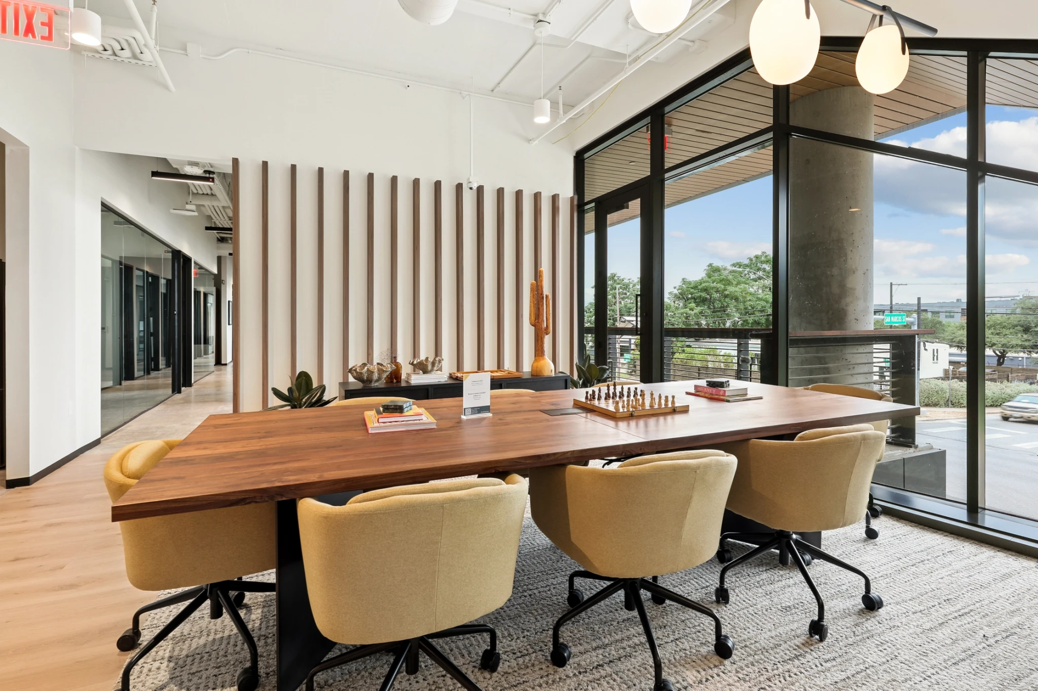 A modern coworking meeting room with a wooden table, six beige chairs, stylish decor, and large windows offering views of the street and trees.