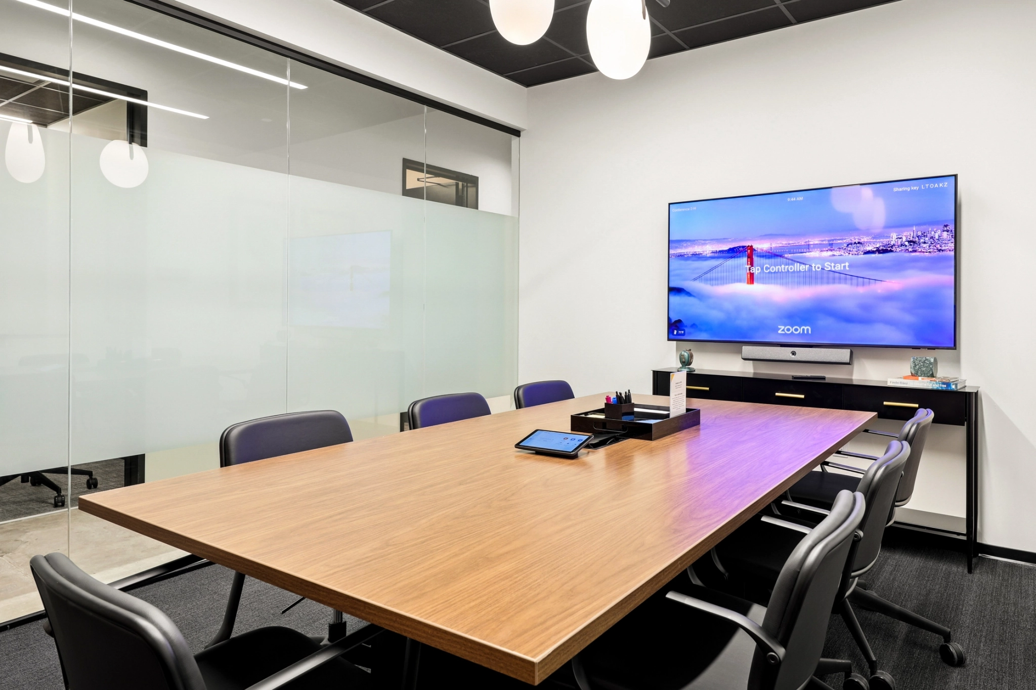 Moderna sala de reuniones con una larga mesa de madera, ocho sillas y un monitor de pared con pantalla zoom. Las paredes de cristal rodean el espacio de trabajo y el material de oficina está cuidadosamente dispuesto sobre la mesa para que las reuniones sean productivas.