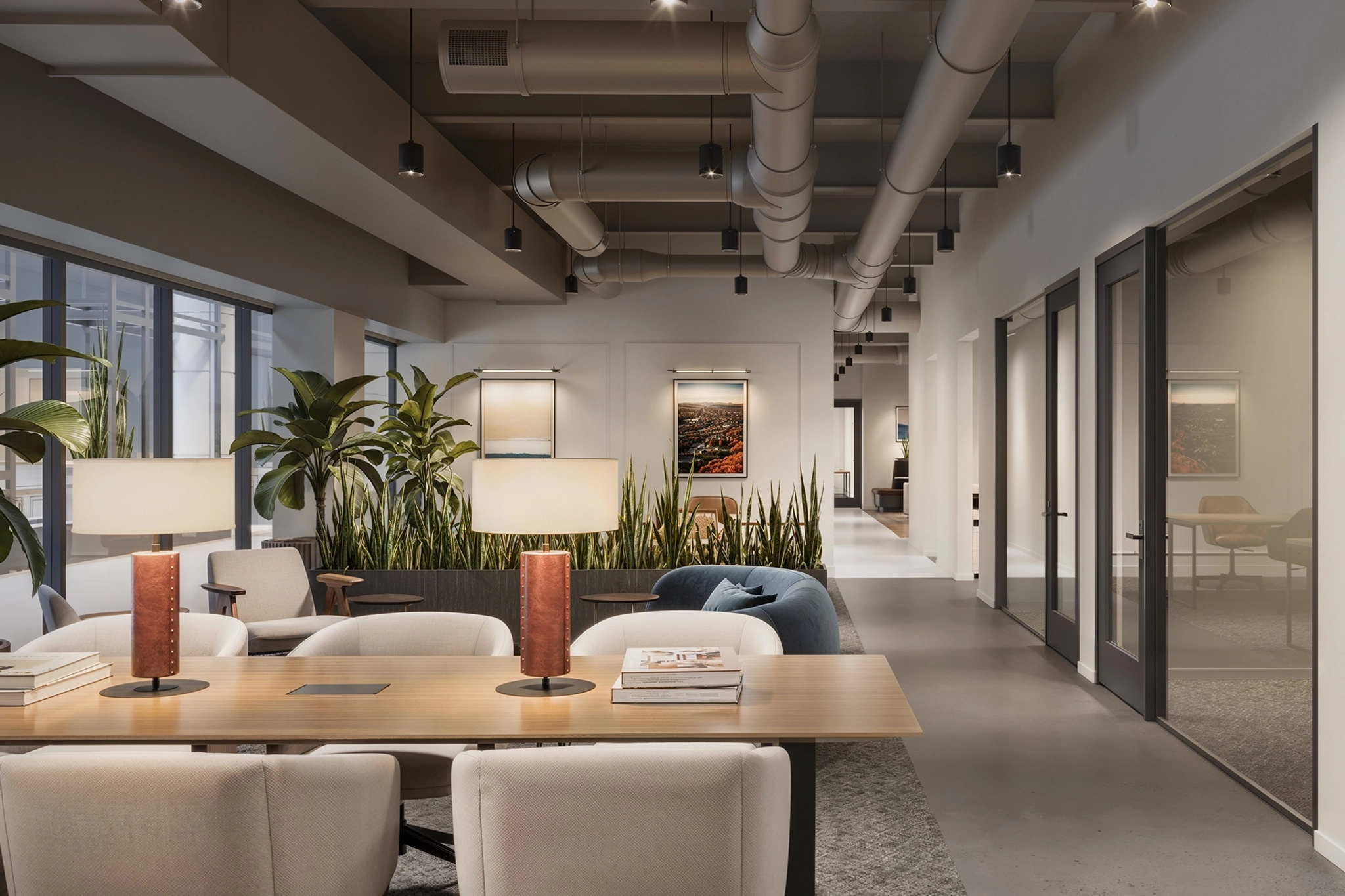 This modern Phoenix coworking space features sleek furniture, pendant lighting, and large windows. A long wooden table with two lamps takes center stage, surrounded by green plants and framed art that adorn the walls, creating an inspiring workspace.