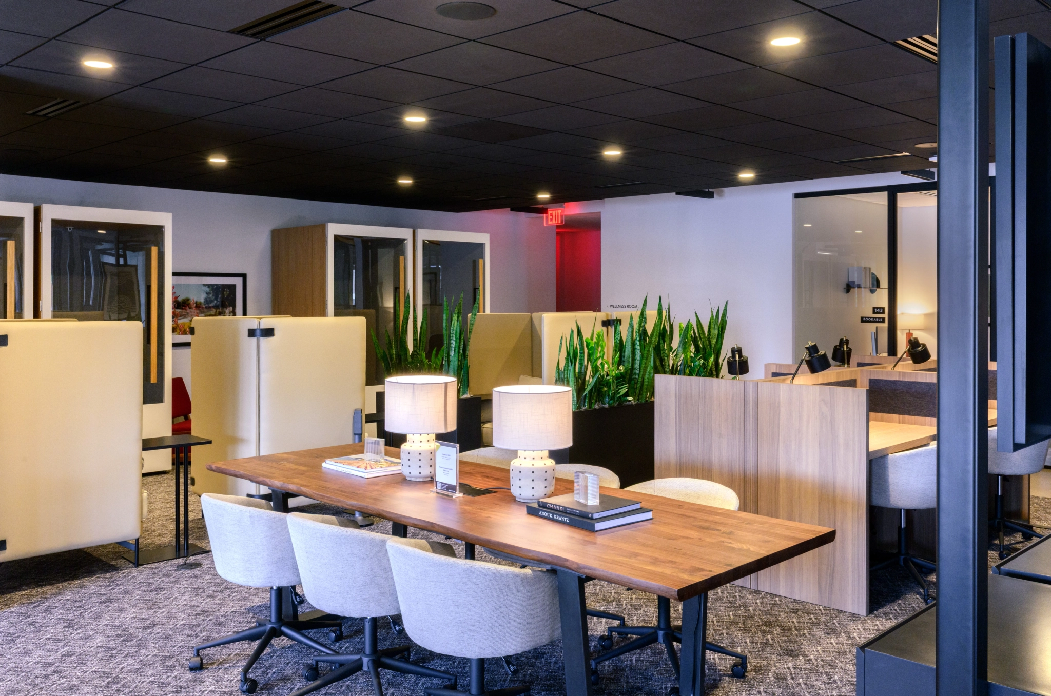 Espace de coworking moderne à Phoenix comprenant une grande table en bois, des chaises, des lampes de bureau, des postes de travail individuels, des plantes d'intérieur et des panneaux de séparation sous un plafond noir épuré.
