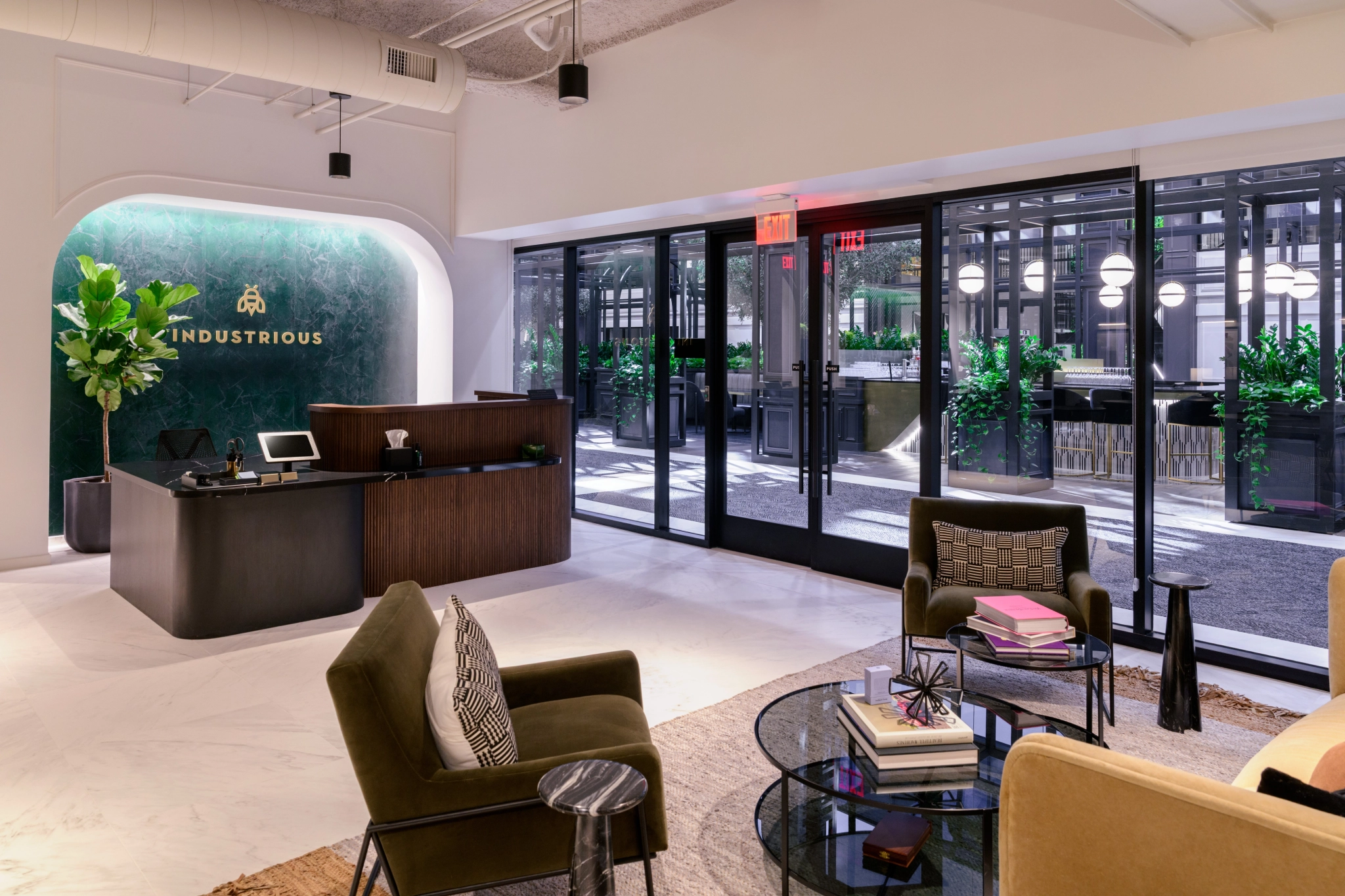 Modern office lobby featuring a reception desk, comfortable seating area, large glass doors, indoor plants, and "Industrious" branding on a green marble wall—ideal for welcoming guests to your workspace.