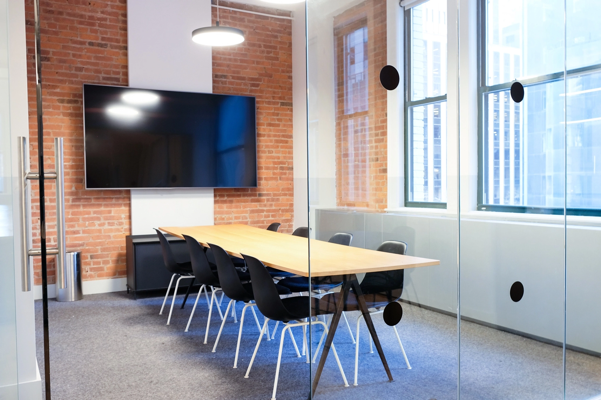 Een moderne vergaderruimte in een coworkingruimte in New York beschikt over een lange houten tafel, acht zwarte stoelen, een grote televisie aan de muur, bakstenen muren, grote ramen en glazen deuren.