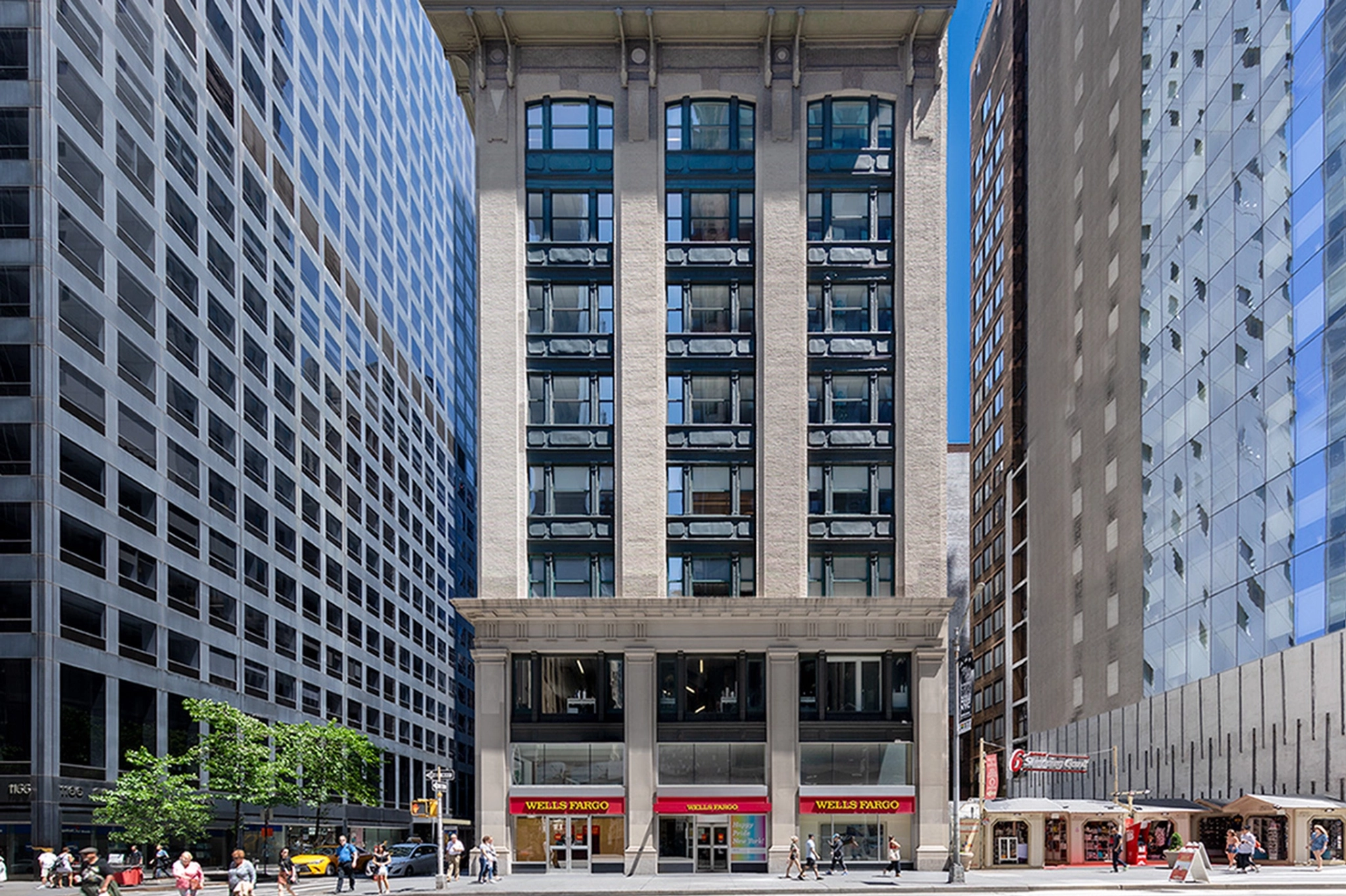 Un immeuble à plusieurs étages avec une banque Wells Fargo au rez-de-chaussée, flanqué de grands immeubles modernes de chaque côté. Plusieurs piétons se trouvent sur le trottoir et certains traversent la rue en direction des bureaux à proximité.