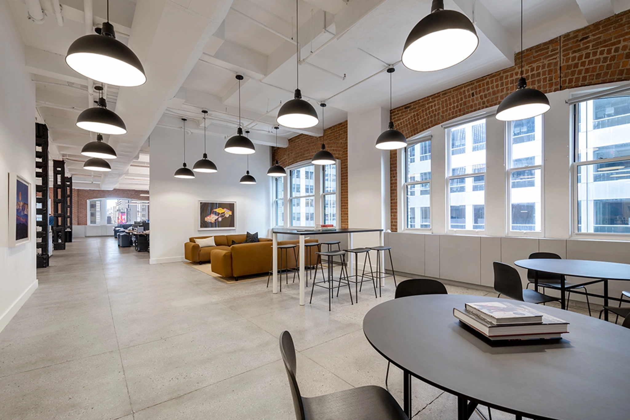 Modern coworking space with large windows, several hanging lights, a lounge area with sofas, round tables with chairs, and a high table with stools. Brick walls and exposed ceiling add to the decor.