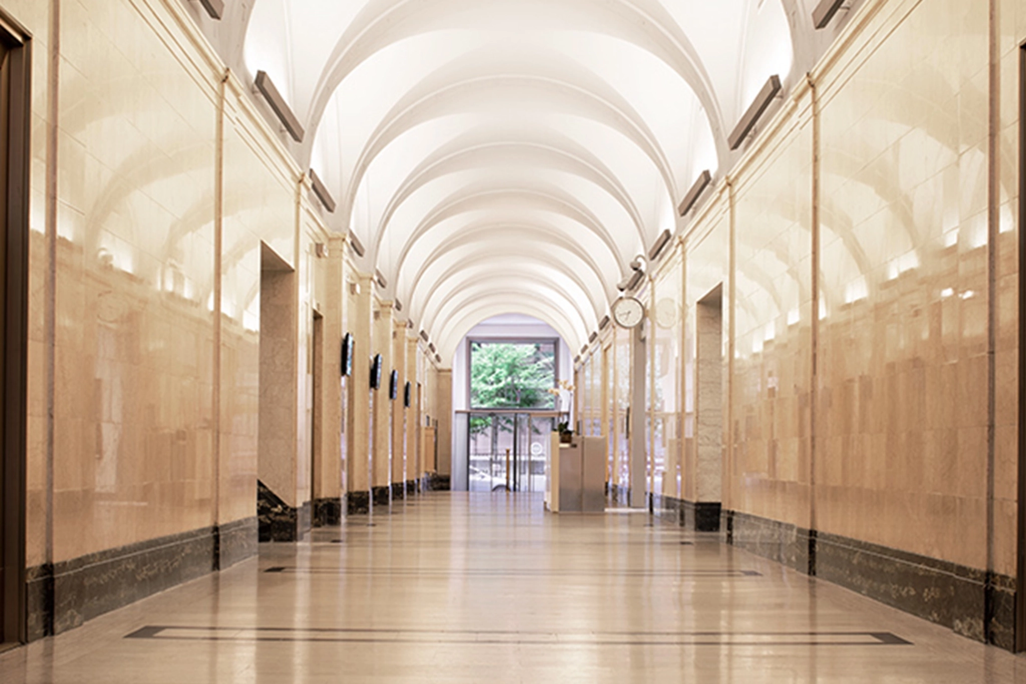 A clean, spacious office hallway with a high arched ceiling and marble walls, leading to a glass door with a view of New York trees outside. A wall clock is mounted near the door, adding a punctual touch to this modern coworking space.