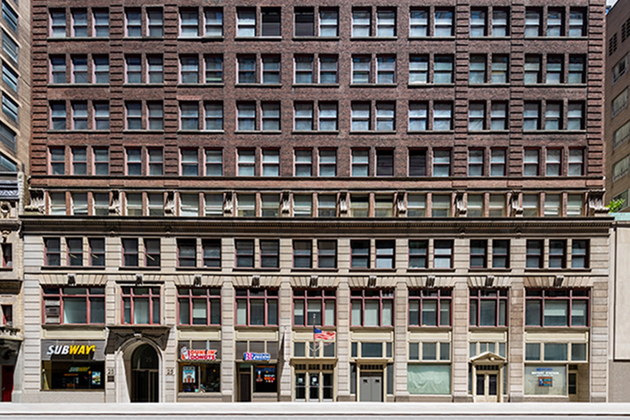 Vooraanzicht van een gebouw met meerdere verdiepingen in New York City met verschillende bedrijven op de begane grond, waaronder Subway, Wingstop en Dunkin'. De bovenste verdiepingen hebben veel ramen en worden gebruikt als coworking-ruimtes.