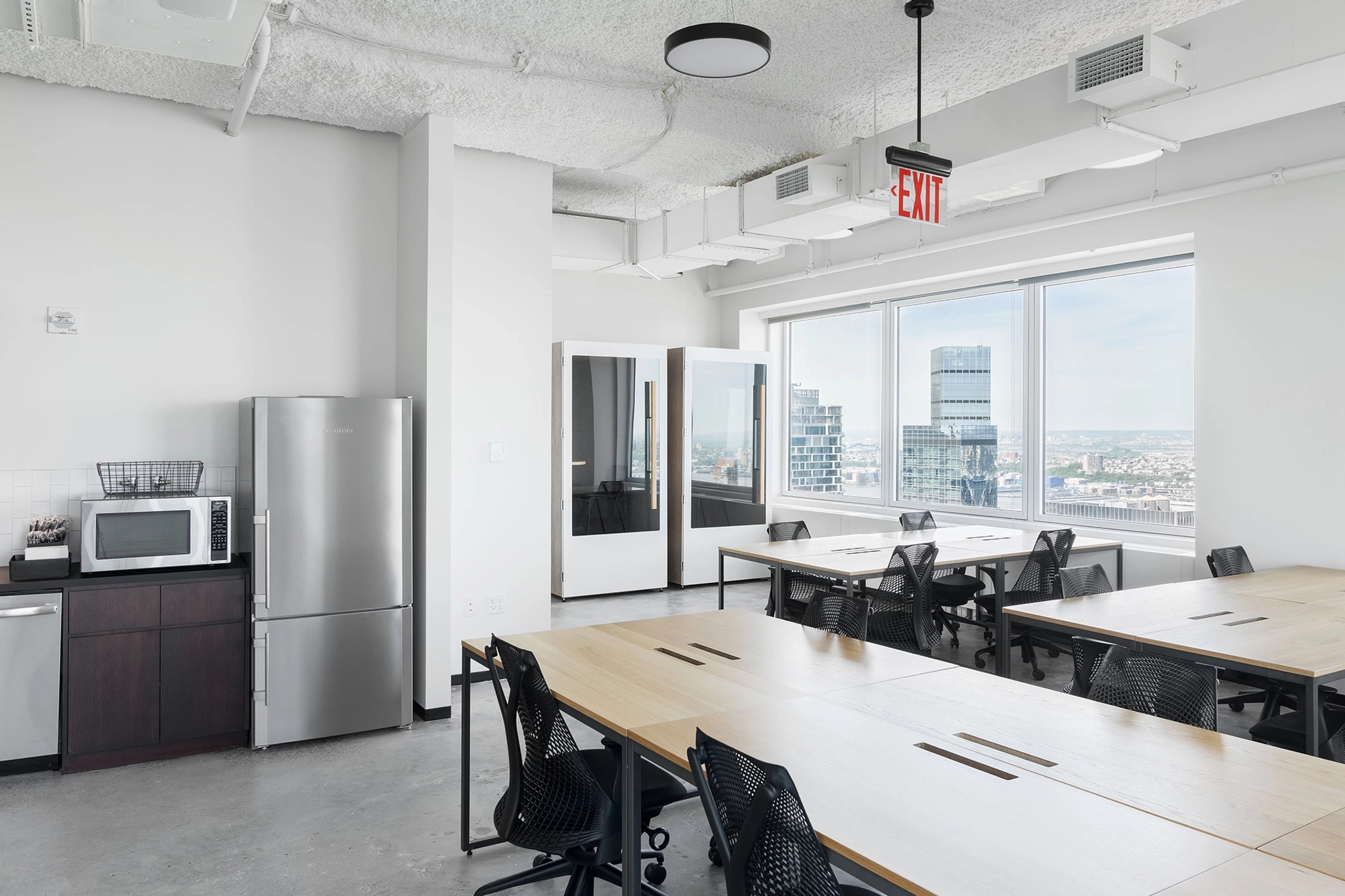 An office break room with tables and chairs, a refrigerator, microwave, and panoramic city views from large windows creates an inviting coworking space.