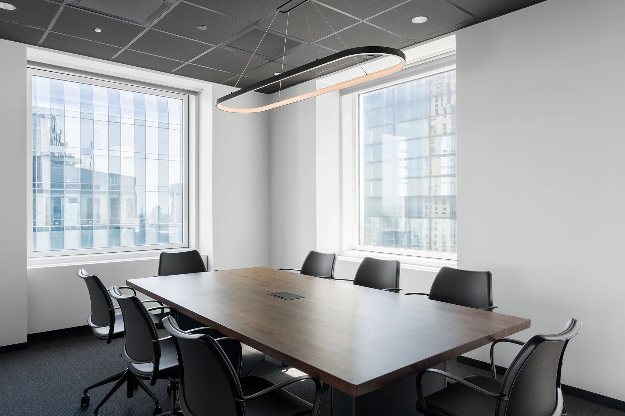 Een moderne coworking-conferentieruimte is voorzien van een lange houten tafel met acht zwarte stoelen, grote ramen met uitzicht op de stad, witte muren en een eigentijdse rechthoekige plafondlamp.