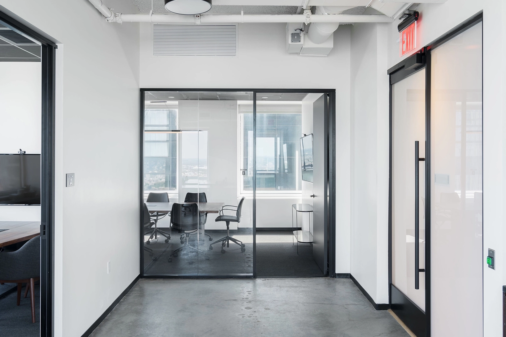 Een moderne kantoorgang in New York City beschikt aan de linkerkant over een met glas omsloten vergaderruimte, uitgerust met een tafel, stoelen en een televisiescherm. Aan de rechterkant wordt een uitgangsdeur aangegeven door een verlicht bord.