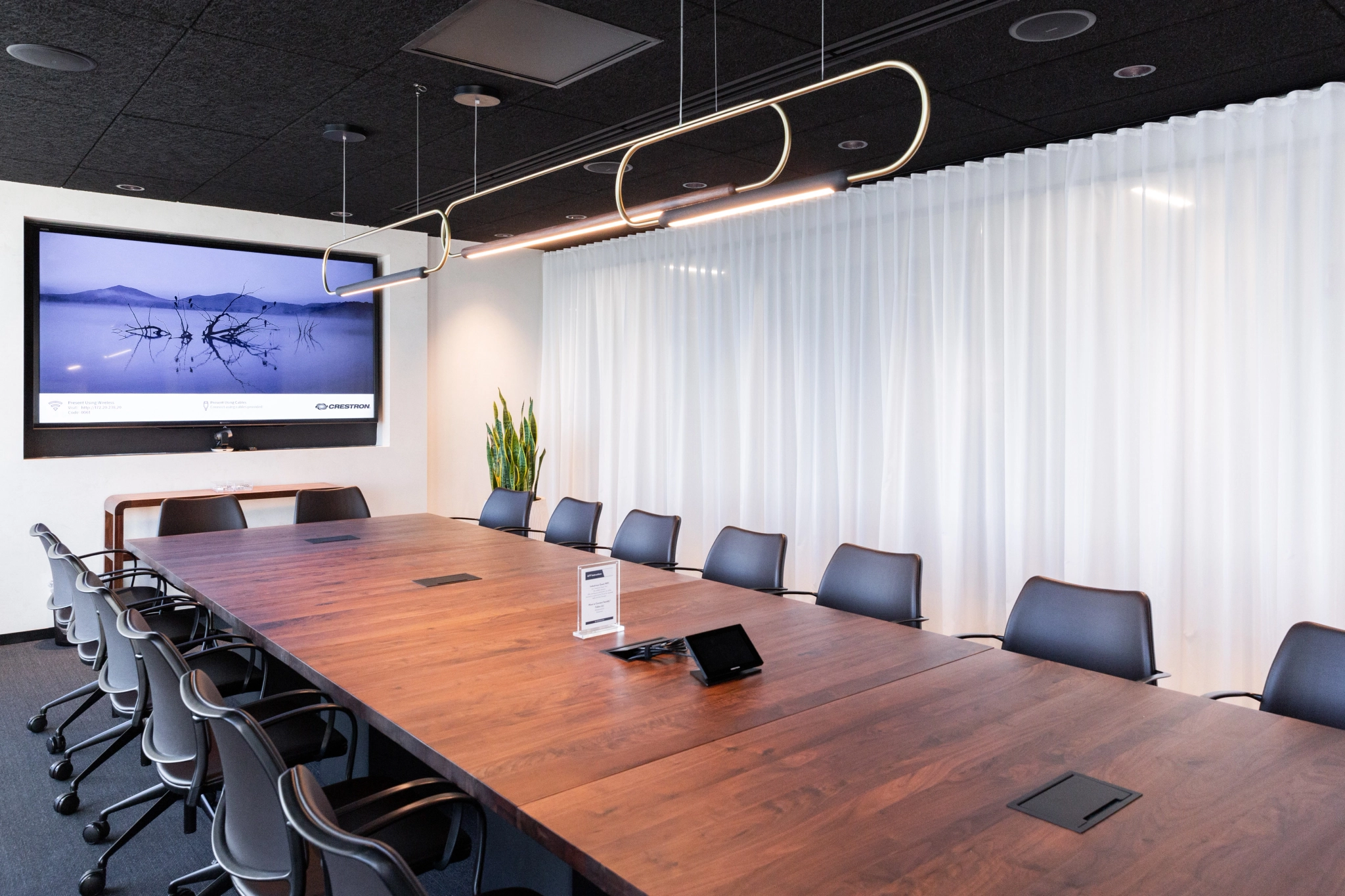 A contemporary meeting room features a long wooden table, several chairs, and a digital screen on the wall, perfect for brainstorming sessions. Hanging lights illuminate the modern workspace while a plant adds a touch of nature.