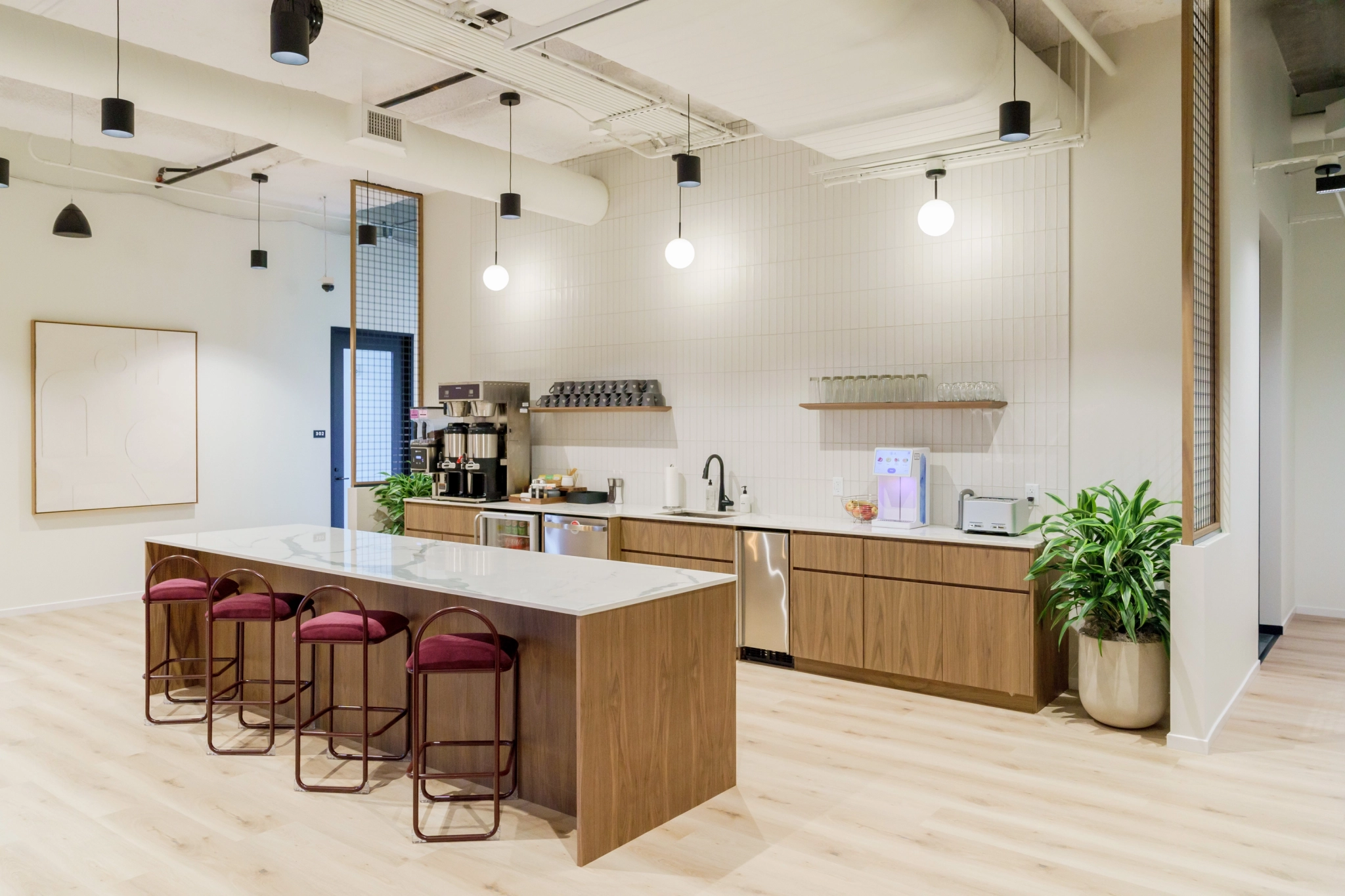 Modern Nashville coworking office kitchen featuring wooden cabinets, a marble-topped island with chairs, lush plants, a sleek coffee machine, and a microwave—all brought together with minimalistic decor for a stylish and functional space.