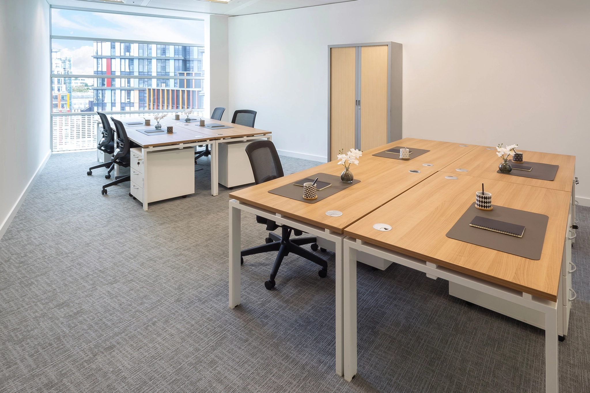 Modern coworking office space with wooden desks, black chairs, and minimal decor. Large window with a city view of London, grey carpet, and a storage cabinet in the background.