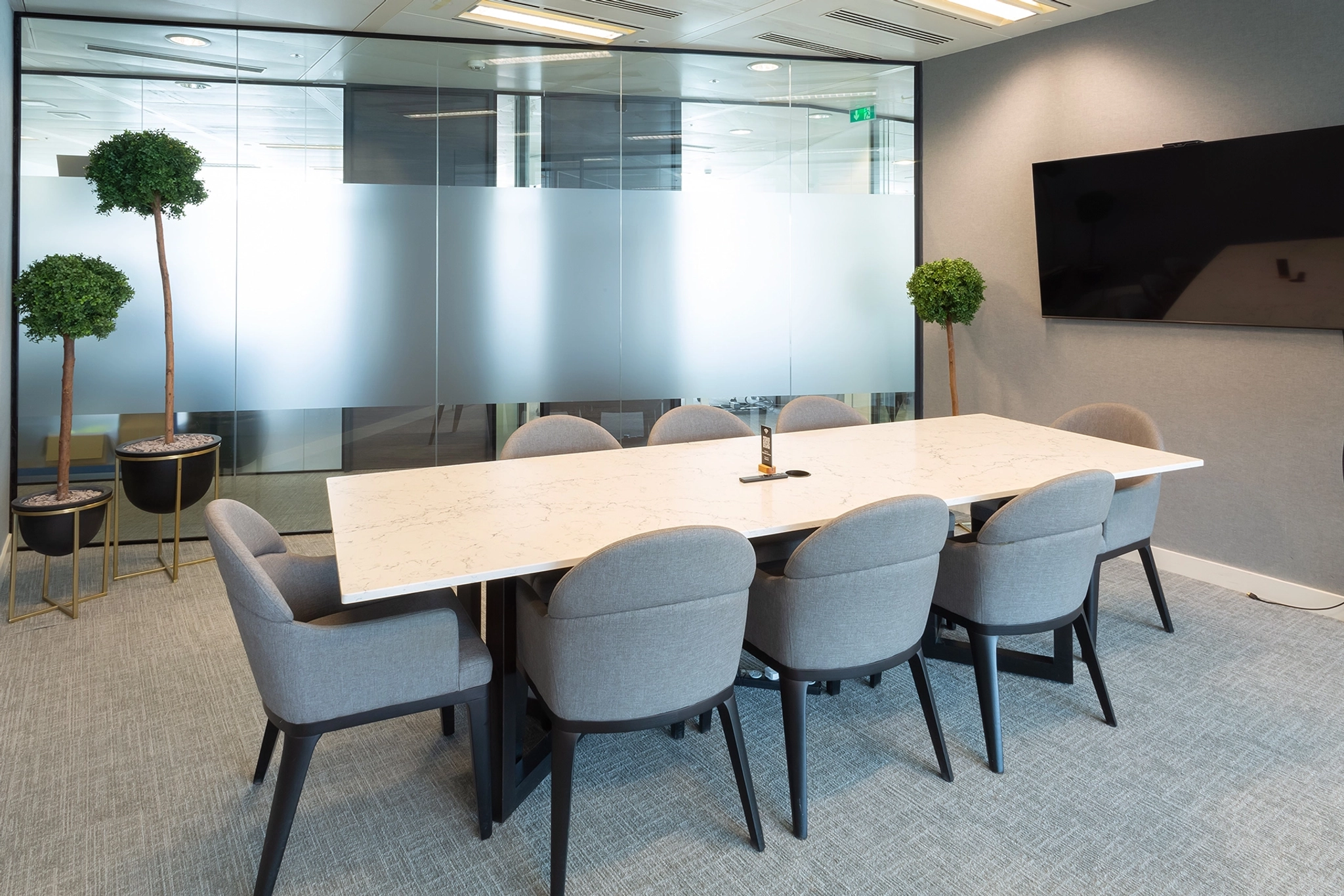 Une salle de réunion moderne dans un bureau londonien comprenant une grande table en marbre blanc, huit chaises grises, une cloison murale en verre, une télévision murale et des plantes en pot.