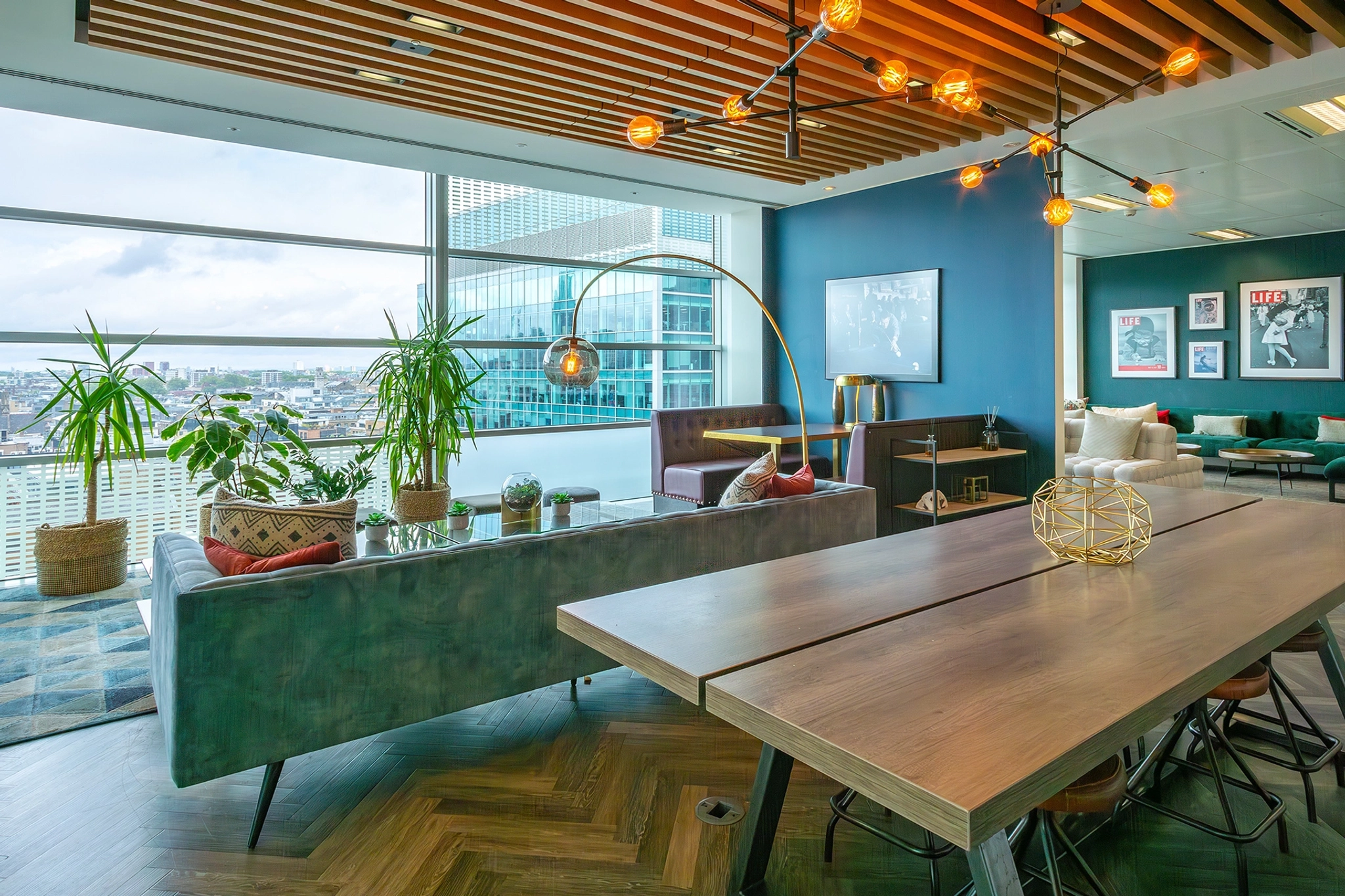 Salon de bureau de coworking moderne avec de grandes fenêtres, des coins salons, une table en bois, des plantes décoratives et des œuvres d'art murales. La pièce est bien éclairée avec des luminaires contemporains et offre une vue sur le paysage urbain, ce qui en fait un espace de travail idéal pour les professionnels.