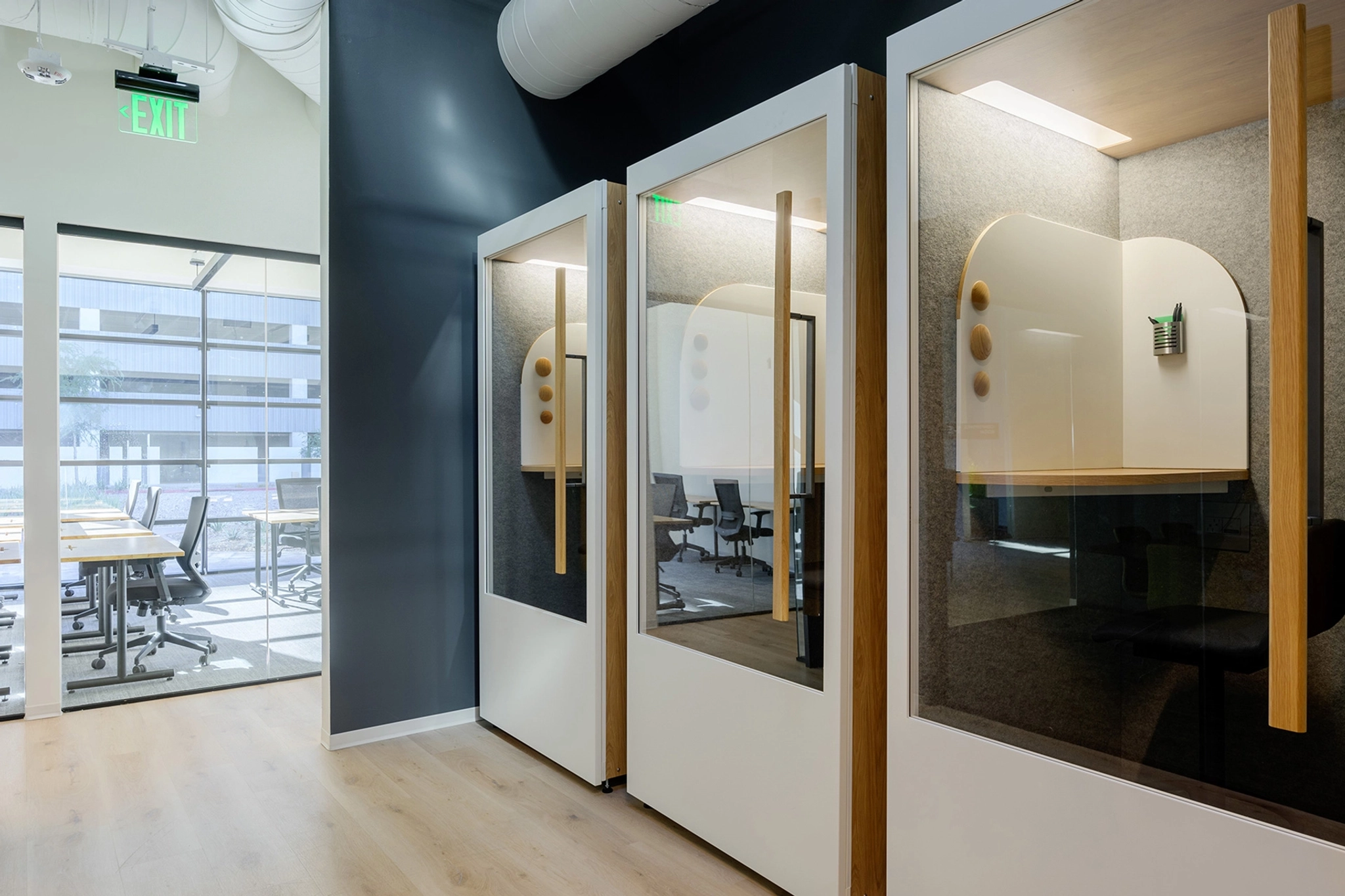 Modern office hallway featuring glass pods with wooden accents for private workspaces reminiscent of Boston's sleek architectural style. A conference room with large windows is visible in the background.