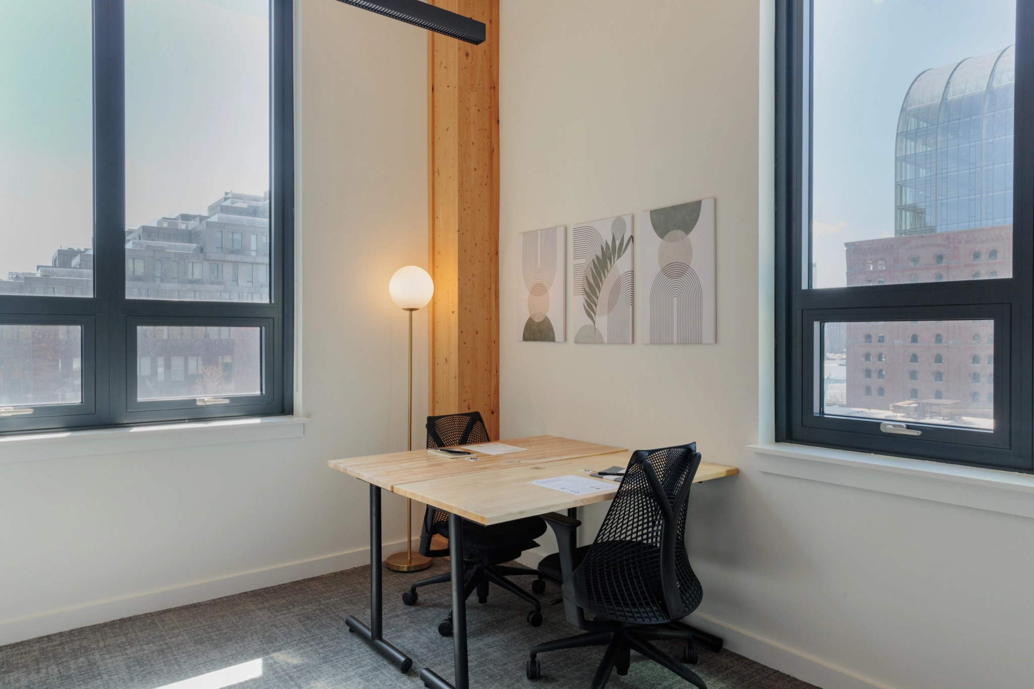 Ein kleines Büro mit zwei modernen Stühlen, einem Holzschreibtisch voller Papiere und großen Fenstern, die natürliches Licht hereinlassen. In der Ecke steht eine Stehlampe und zwischen den Fenstern hängen Kunstwerke, die einen idealen Arbeitsplatz schaffen.