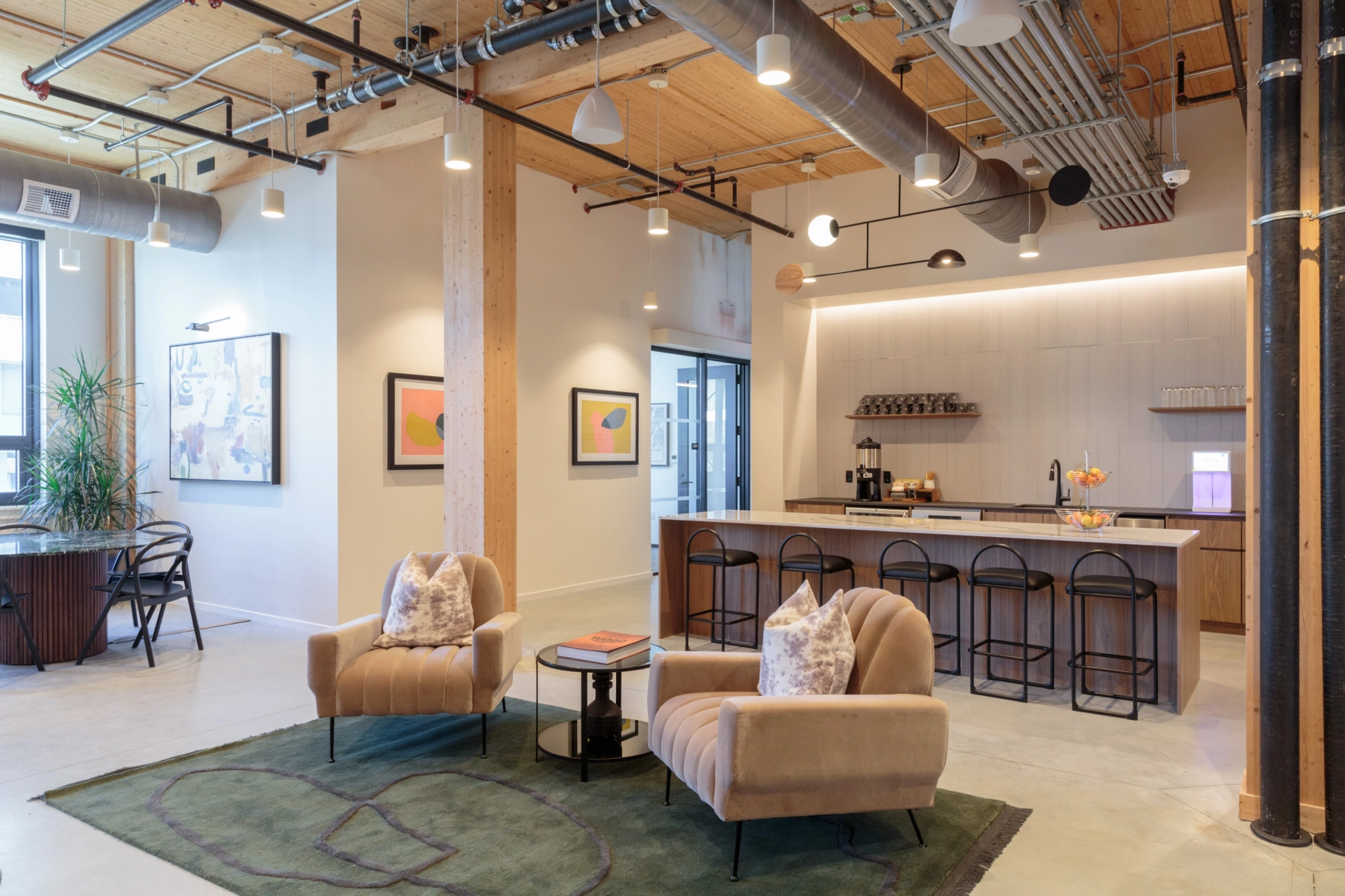 A modern office lounge with two beige chairs and a circular coffee table on a green rug. In the background, there's a bar area with stools and a kitchenette under linear lighting. Artwork decorates the walls, creating an inviting workspace perfect for informal meetings.