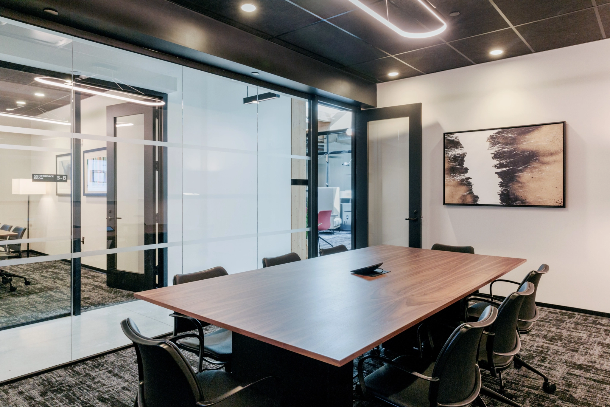 A modern coworking conference room in Boston with a wooden table, eight chairs, a glass wall, and abstract art on one side. The workspace features minimalist decor and essential office equipment.