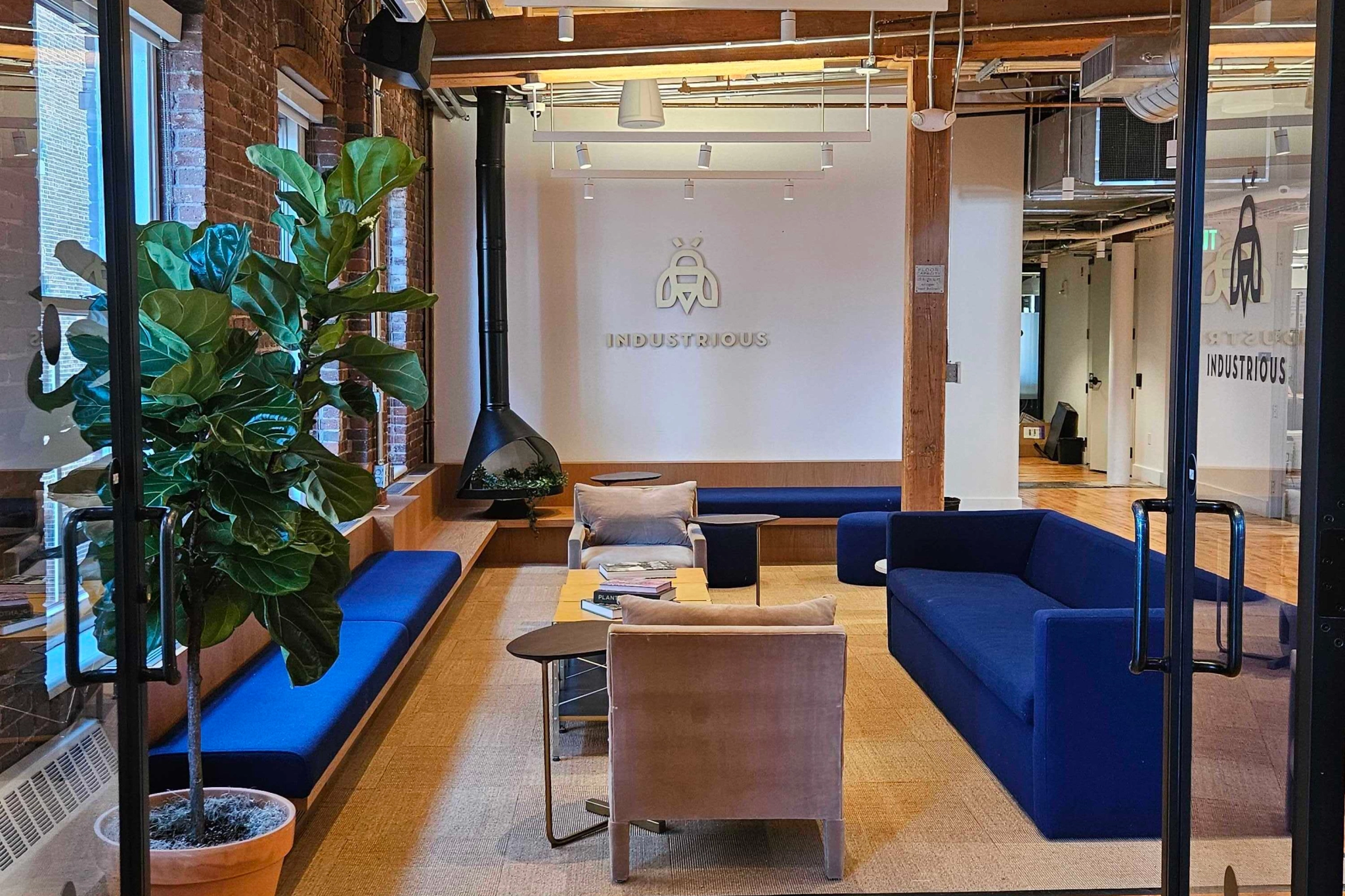 Modern coworking lounge in Boston with navy couches, a light tan armchair, and a large potted plant. "Industrious" logo on the wall enhances the inviting workspace ambiance.