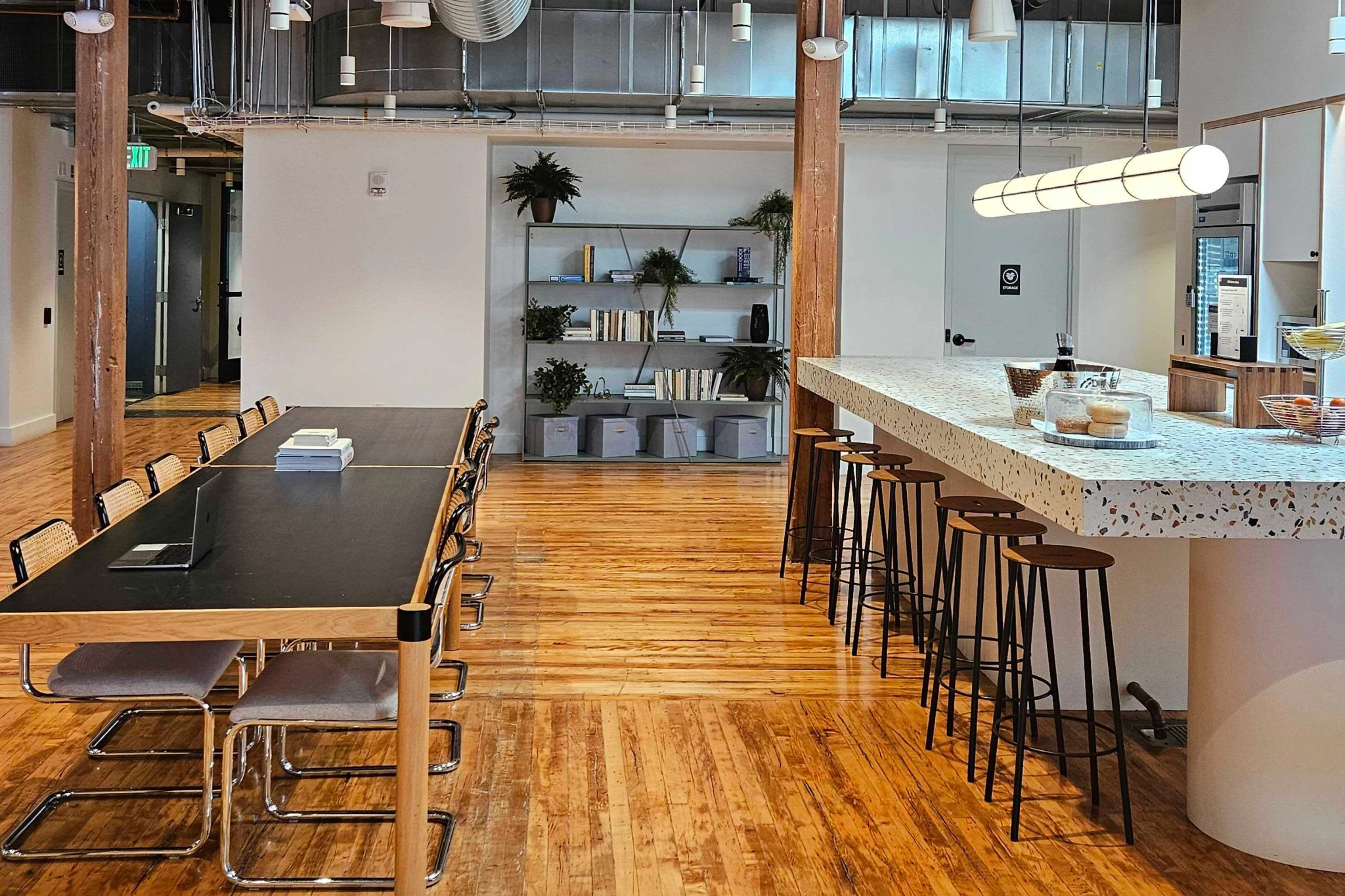 A modern office kitchen in a Boston coworking space features a long black table, chairs, and a bar with stools. Shelves adorned with plants and items provide a stylish background, while the wooden floor adds warmth to this inviting meeting area.