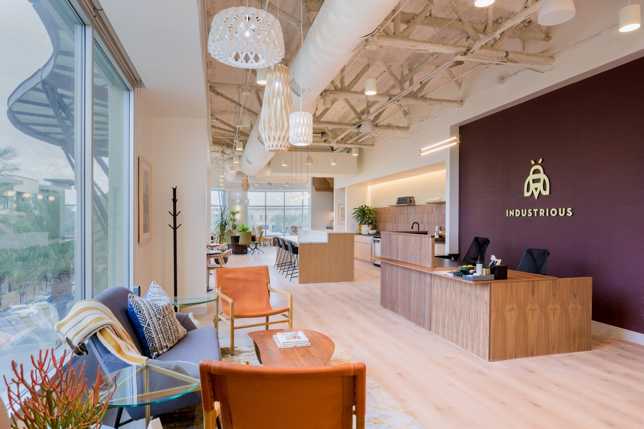 A modern office lobby with a reception desk, lounge seating, and a purple wall displaying the "Industrious" logo. Large windows line the left side, allowing natural light to brighten the coworking space. Perfect for professionals in Austin, it even features a well-equipped meeting room for all your needs.