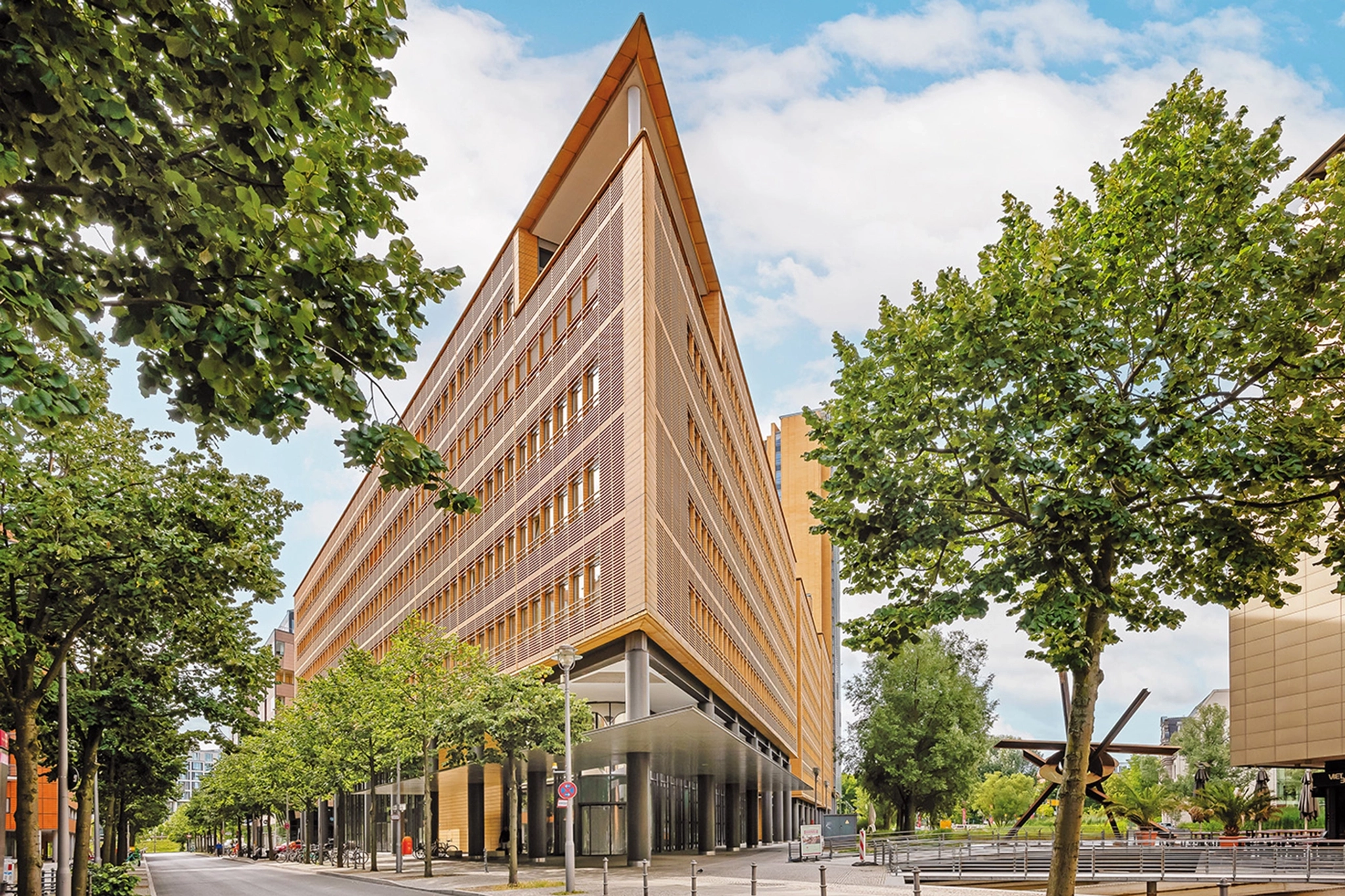 Een modern, driehoekig gebouw van zeven verdiepingen met grote ramen, geflankeerd door bomen aan een Berlijnse stadsstraat, onder een gedeeltelijk bewolkte, helderblauwe hemel, biedt ideale werkruimteoplossingen en vergaderruimtes.