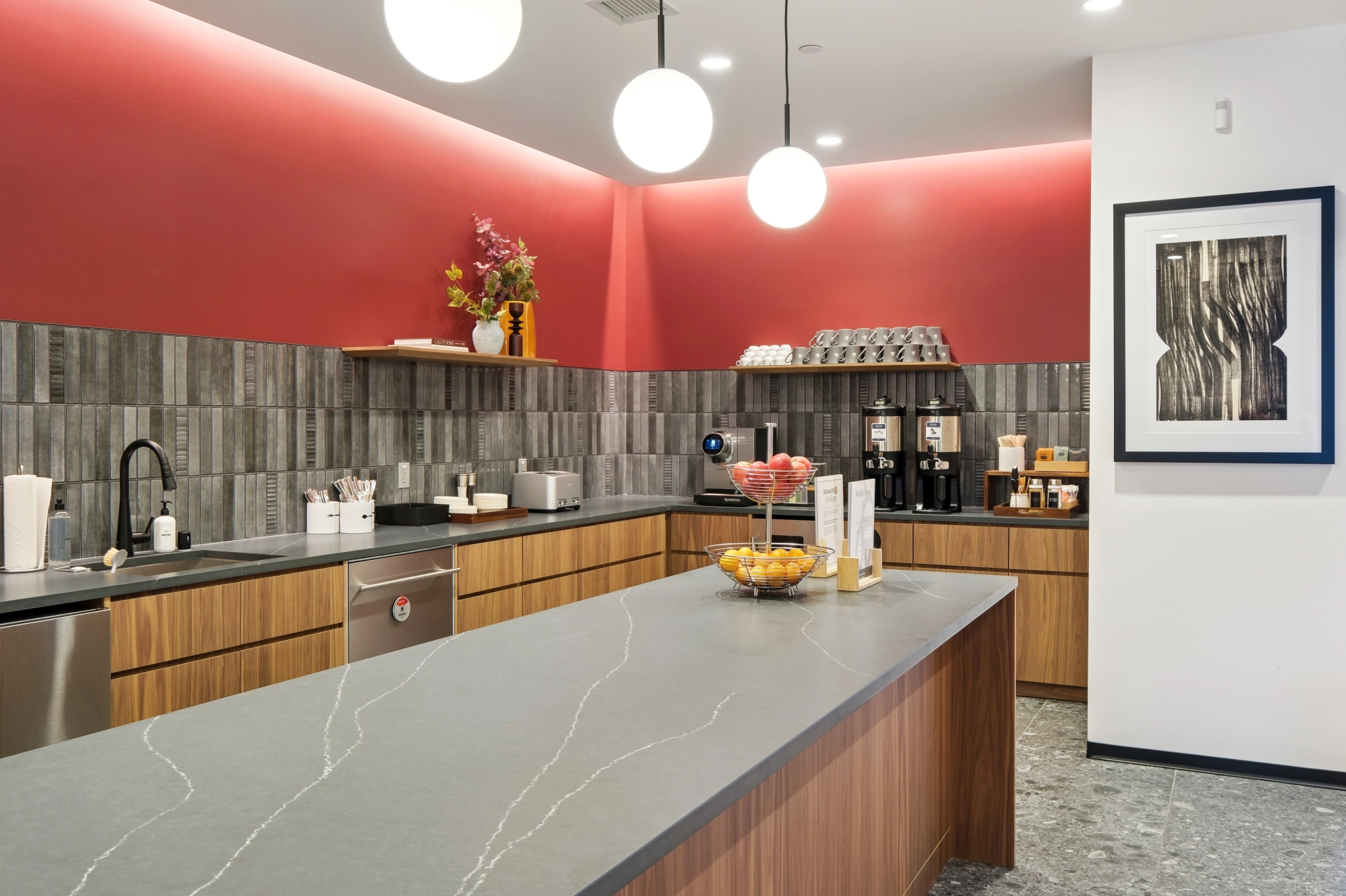 Modern coworking kitchenette with gray countertops, wood cabinets, coffee machines, snack bowls, and wall art, under round ceiling lights and a red accent wall—perfect for breaks in your vibrant workspace.