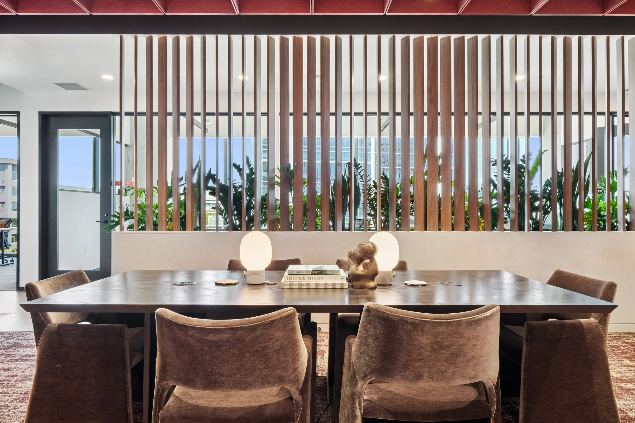 A modern San Diego meeting room with a large wooden table, velvet chairs, two table lamps, decorative objects, and a slatted wood partition in the background.