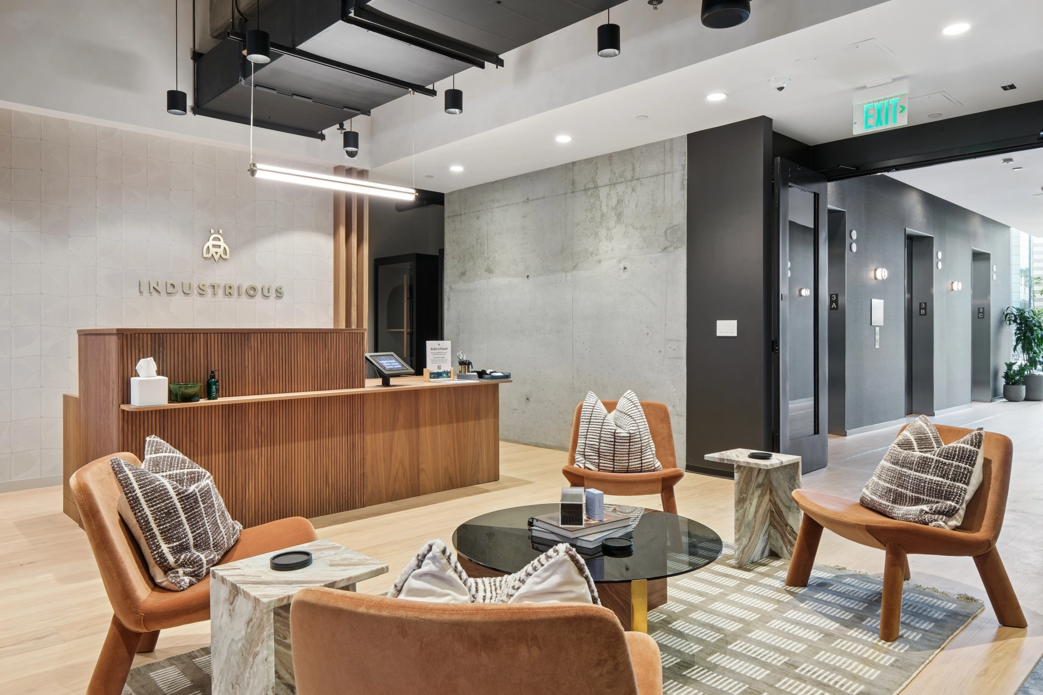 Modern office lobby featuring a wooden reception desk, four brown chairs around a glass coffee table, and neutral-toned decor. The "Industrious" logo is on the back wall, creating an inviting workspace atmosphere.