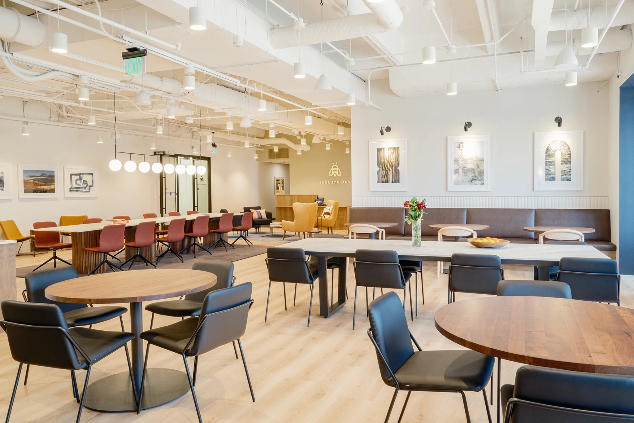 The modern coworking lounge features round tables and black chairs alongside a long meeting table with vibrant red seats. Framed art adorns the walls, while overhead lights and light wooden flooring create a bright atmosphere, perfect for collaborative work in Carmel.