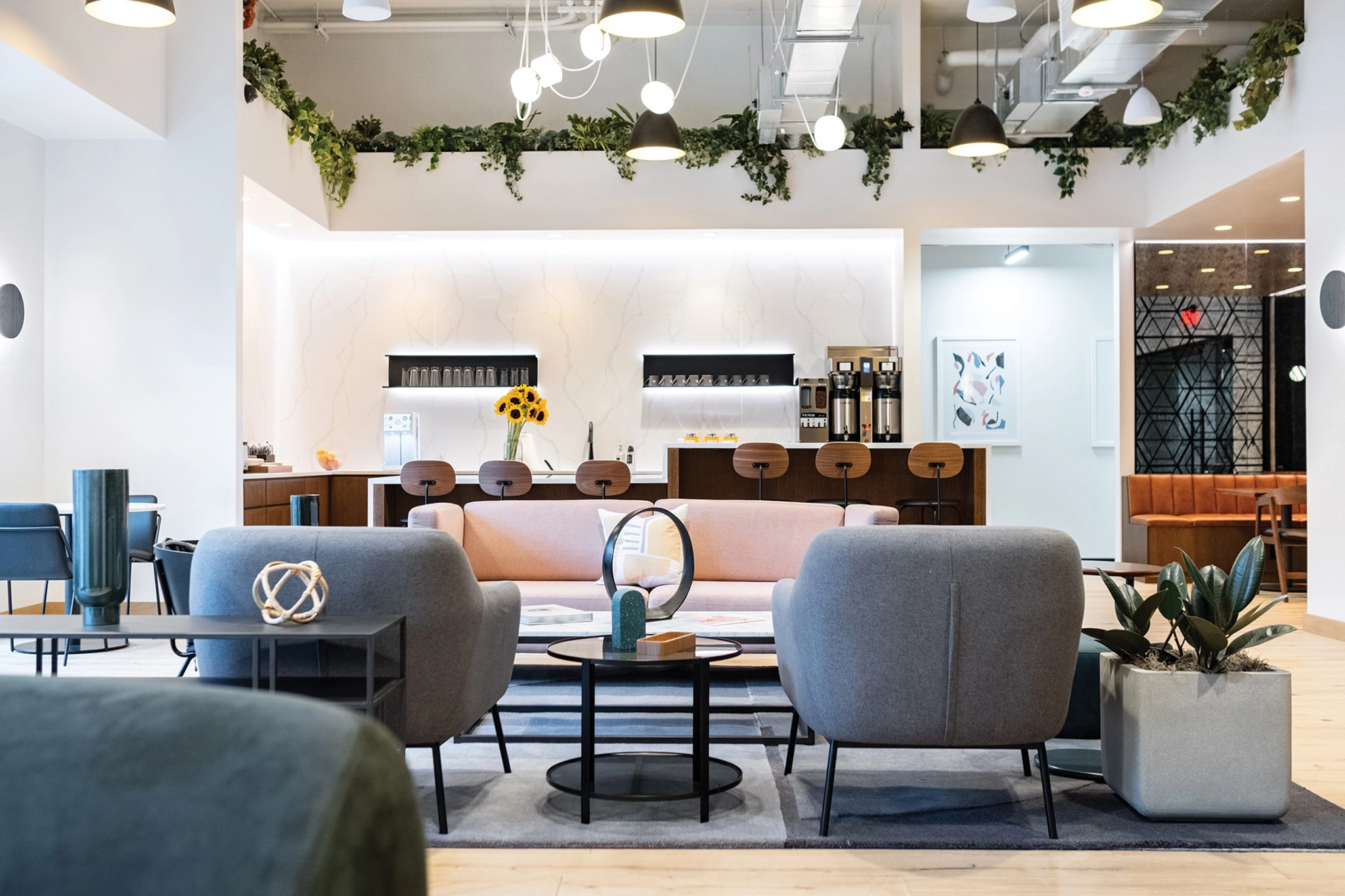 Modern lounge in a vibrant coworking space with gray and pink seating, a central coffee table, and a counter with barstools in the background. Hanging lights and indoor plants enhance the decor, creating an inviting atmosphere perfect for your Phoenix office needs.