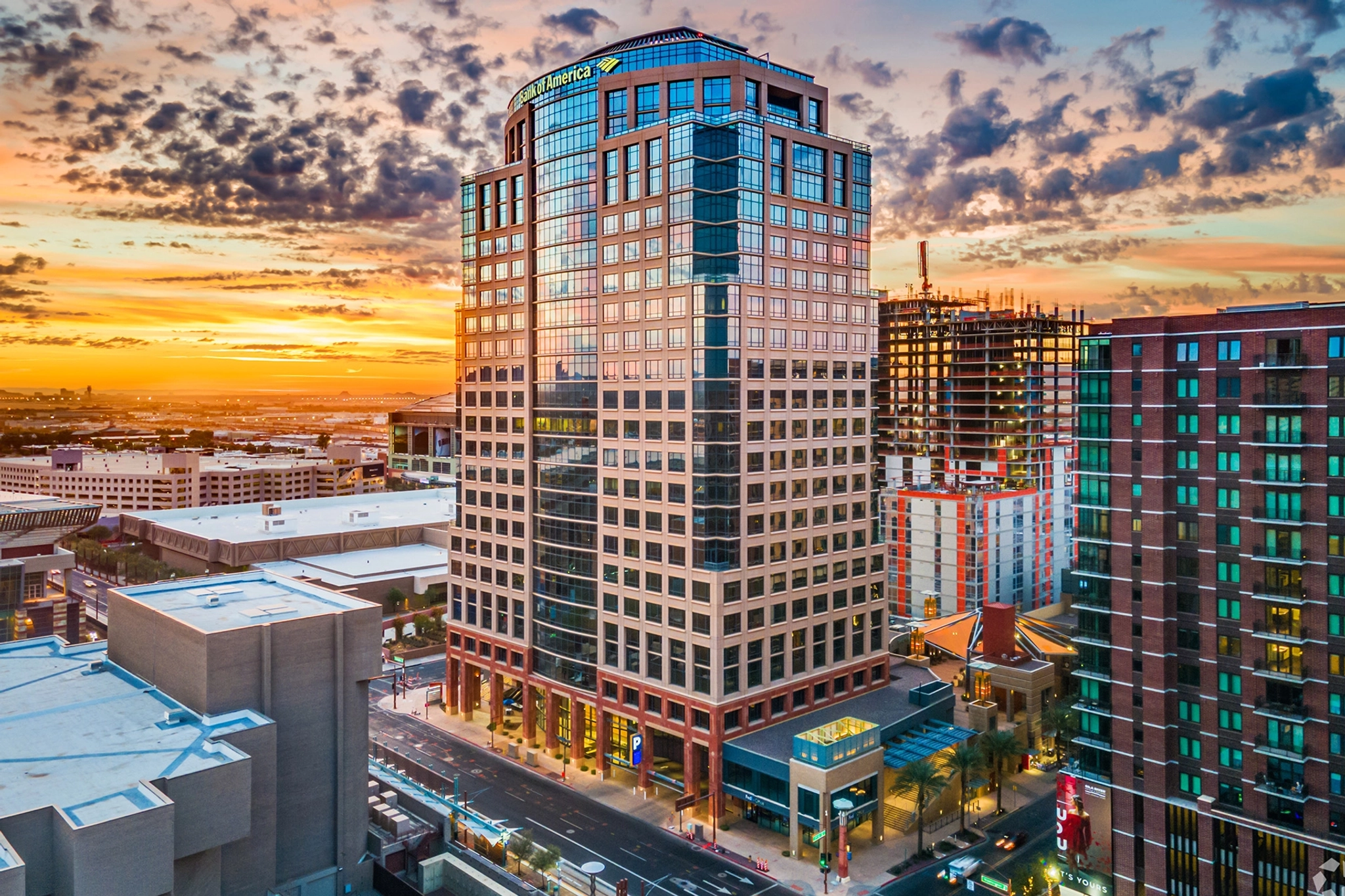 Aerial view of a large office building in a city at sunset with modern coworking spaces surrounded by other buildings and construction sites under a partly cloudy sky.