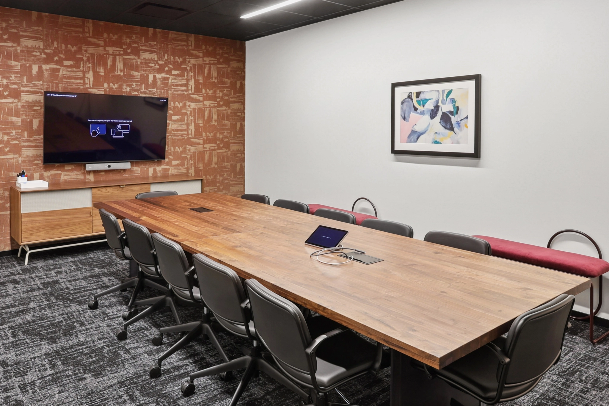 A modern Phoenix workspace conference room with a long wooden table, black chairs, a wall-mounted screen, abstract artwork, and a bench against the wall.