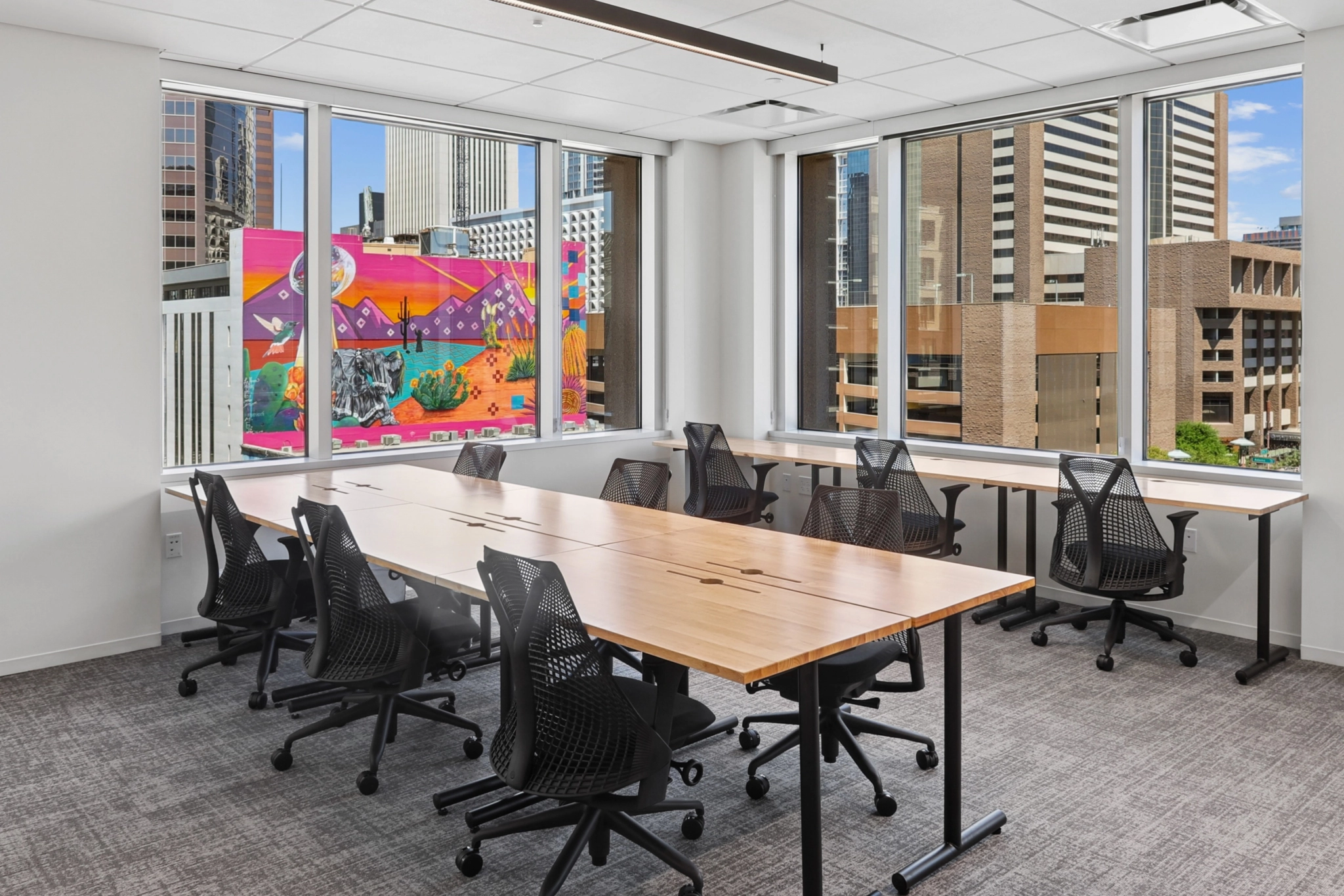 Modern Phoenix office featuring eight black mesh chairs around two wooden tables, large windows with city views, and a colorful mural visible outside—an inspiring workspace for your team.