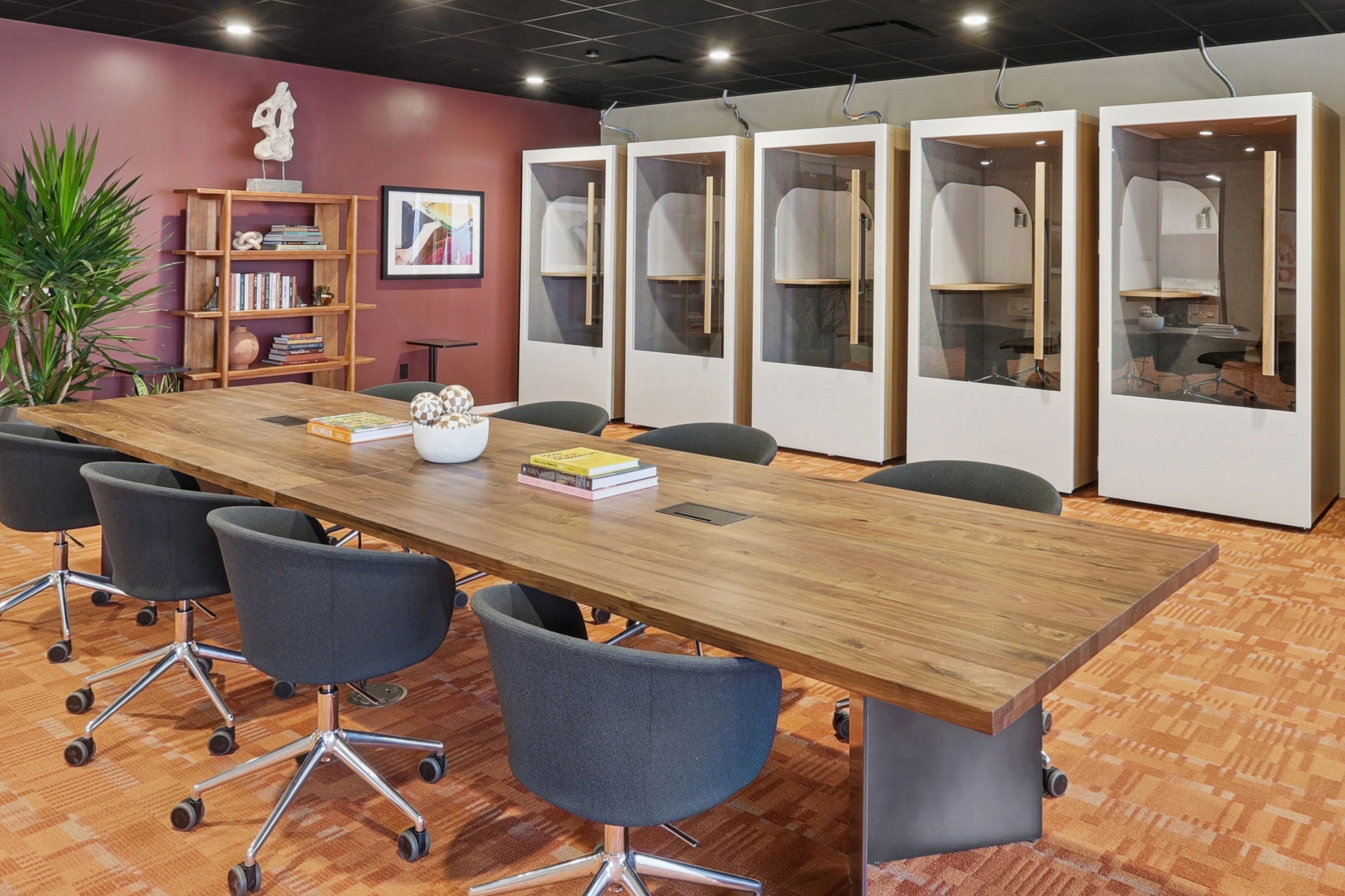 A modern coworking meeting room in Phoenix features a long wooden table, black chairs, bookshelves, a potted plant, and four individual work pods lining the back wall.