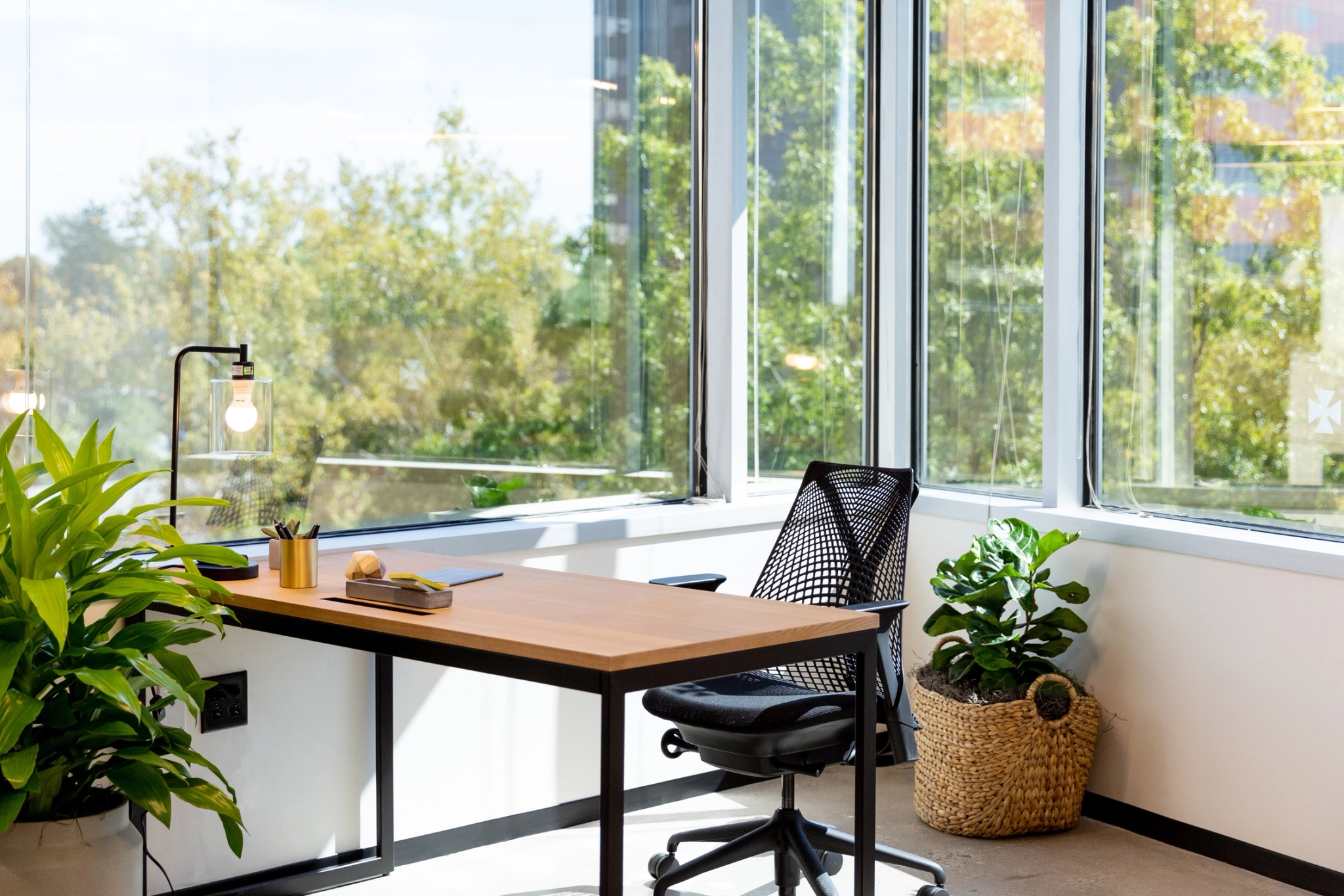 A modern workspace corner features a wooden desk, mesh chair, desk lamp, notepad, pen, and potted plants. Large windows offer a view of trees outside, transforming this office area into an inspiring environment.