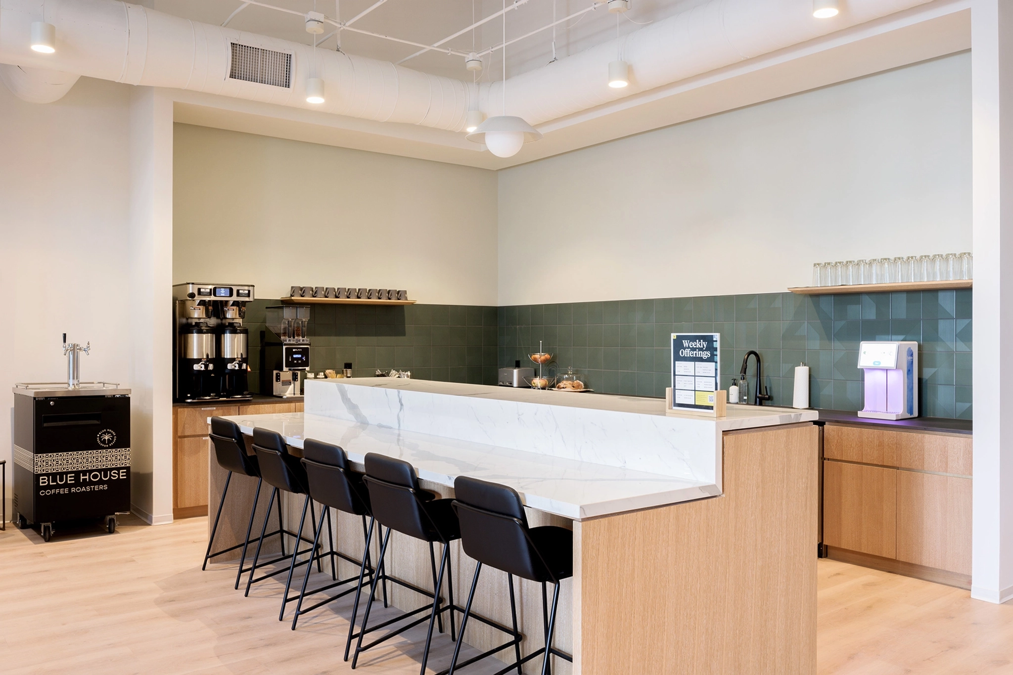 In a bustling Palo Alto coworking space, a modern coffee counter features five barstools, state-of-the-art coffee machines, and a tempting display of pastries. Light-colored walls complement the gray-green backsplash tiles, while ceiling lights warmly illuminate the area.