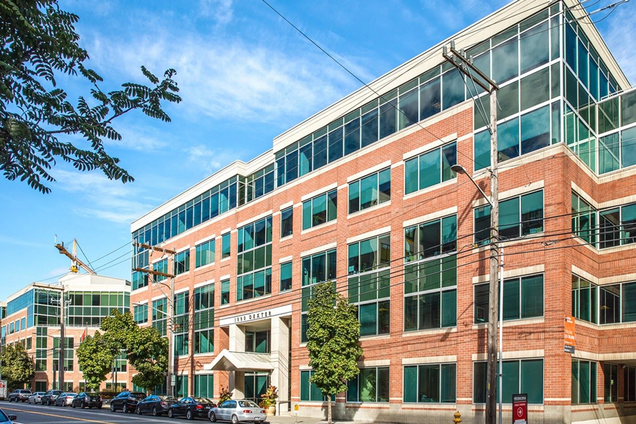 A red brick office building with large windows under a blue sky in Seattle, featuring trees and parked cars in front. This inviting workspace offers a perfect setting for productivity, complete with modern meeting rooms to facilitate collaboration.