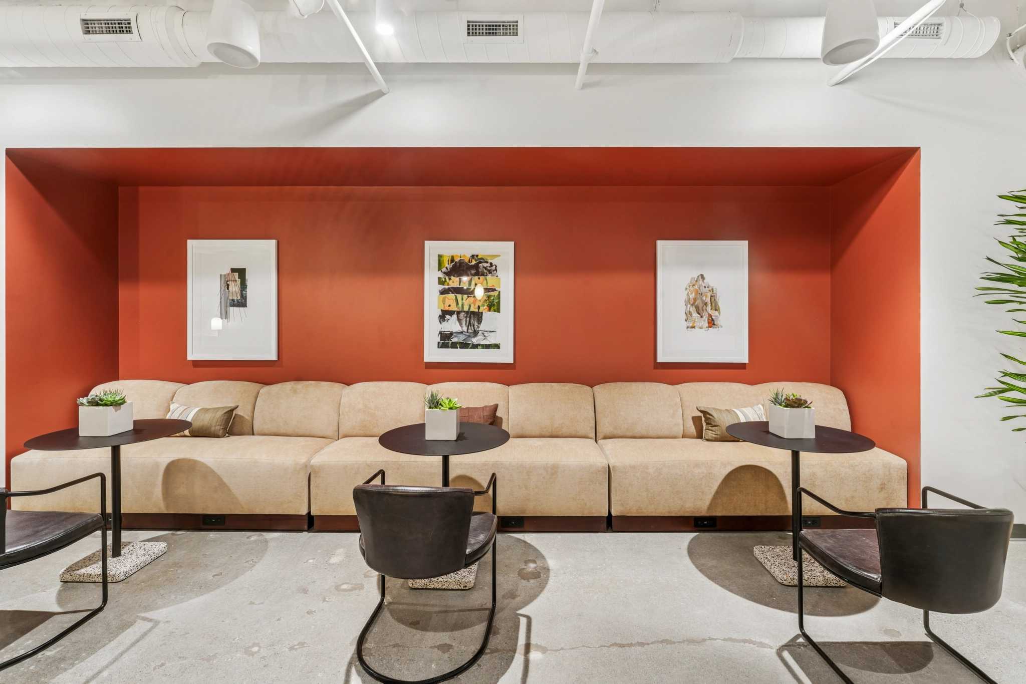 Espace de coworking moderne composé de canapés beiges, de chaises noires, de tables rondes, de petites plantes en pot et de trois œuvres d'art encadrées sur un mur d'accent rouge dans une salle de réunion lumineuse de Seattle.