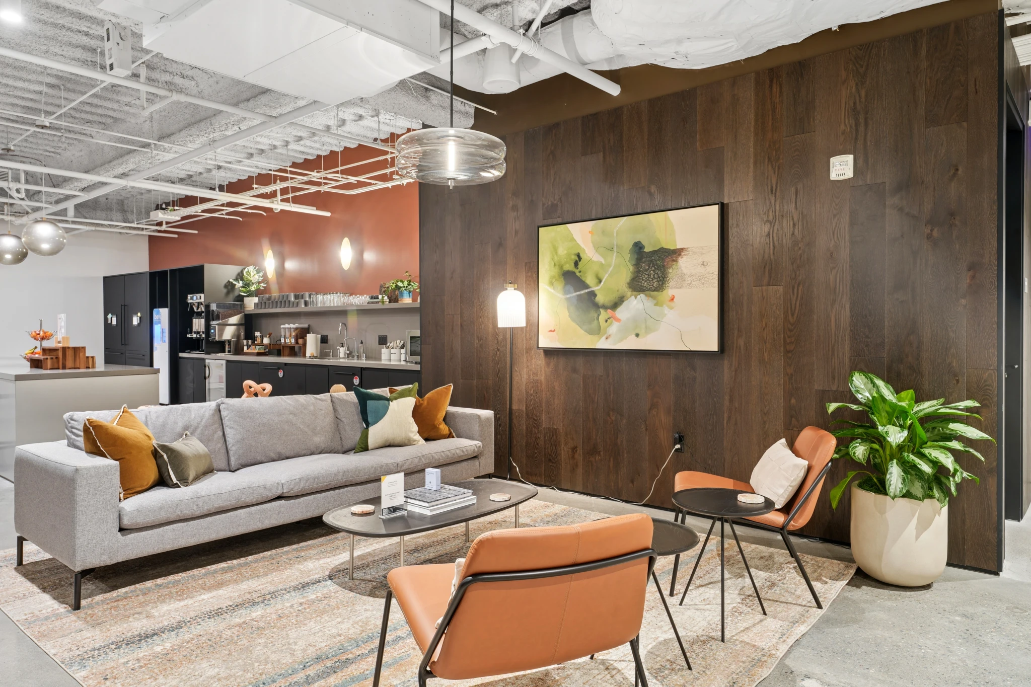 Moderne coworking lounge met een grijze bank, twee oranje stoelen, een salontafel, muurkunst, een potplant en een kitchenette op de achtergrond - perfect voor een uitnodigende werkruimte of informele vergaderruimte.