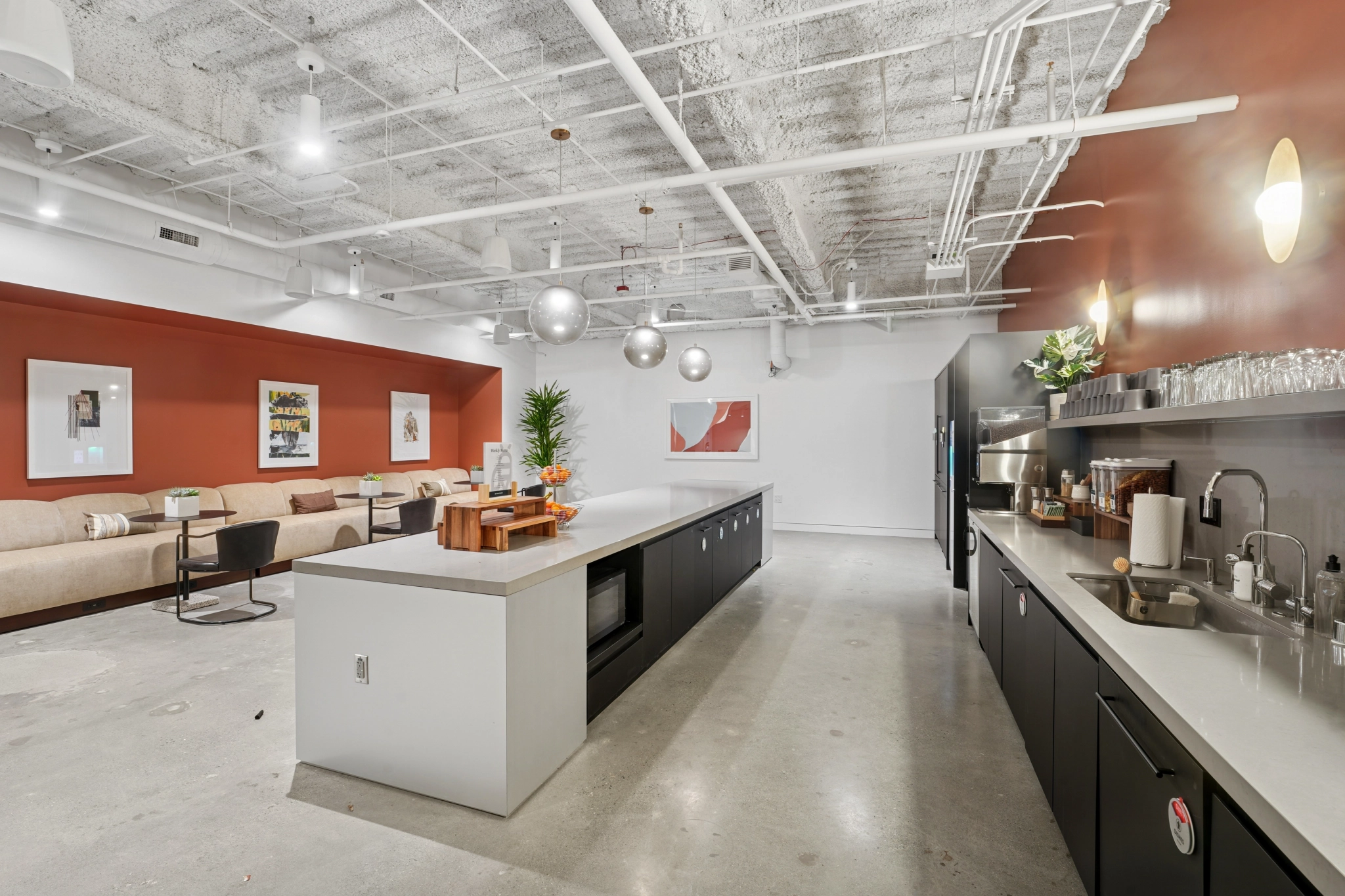 Modern communal kitchen and lounge area in Seattle with a large island, black cabinets, workspace seating, overhead lighting, wall art, and a beige sofa against a burnt orange accent wall.
