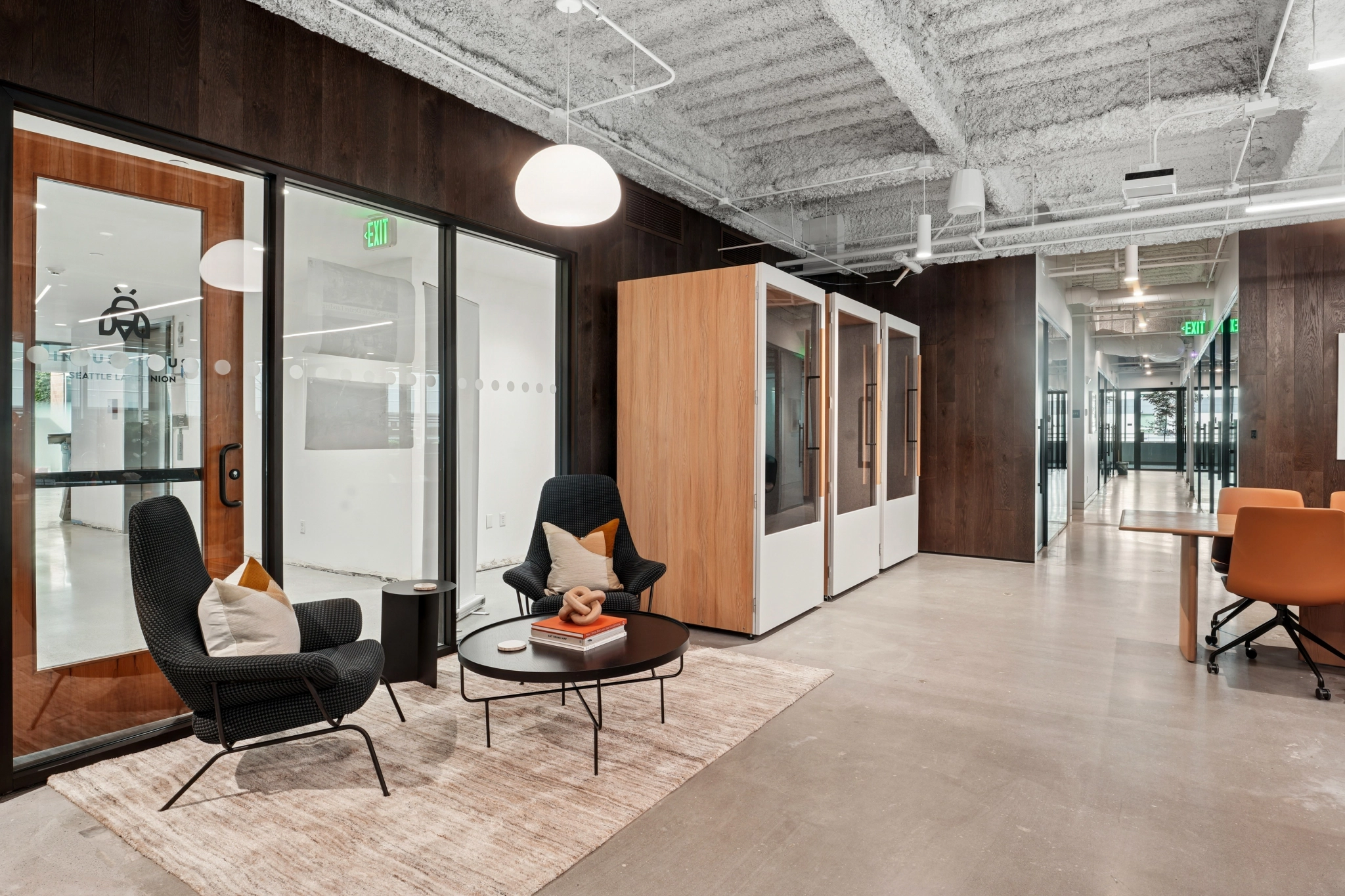 Modern Seattle office lounge area with two black chairs, a small round table, a rug, and private phone booths along the wall. Glass doors and an open office space are visible in the background.