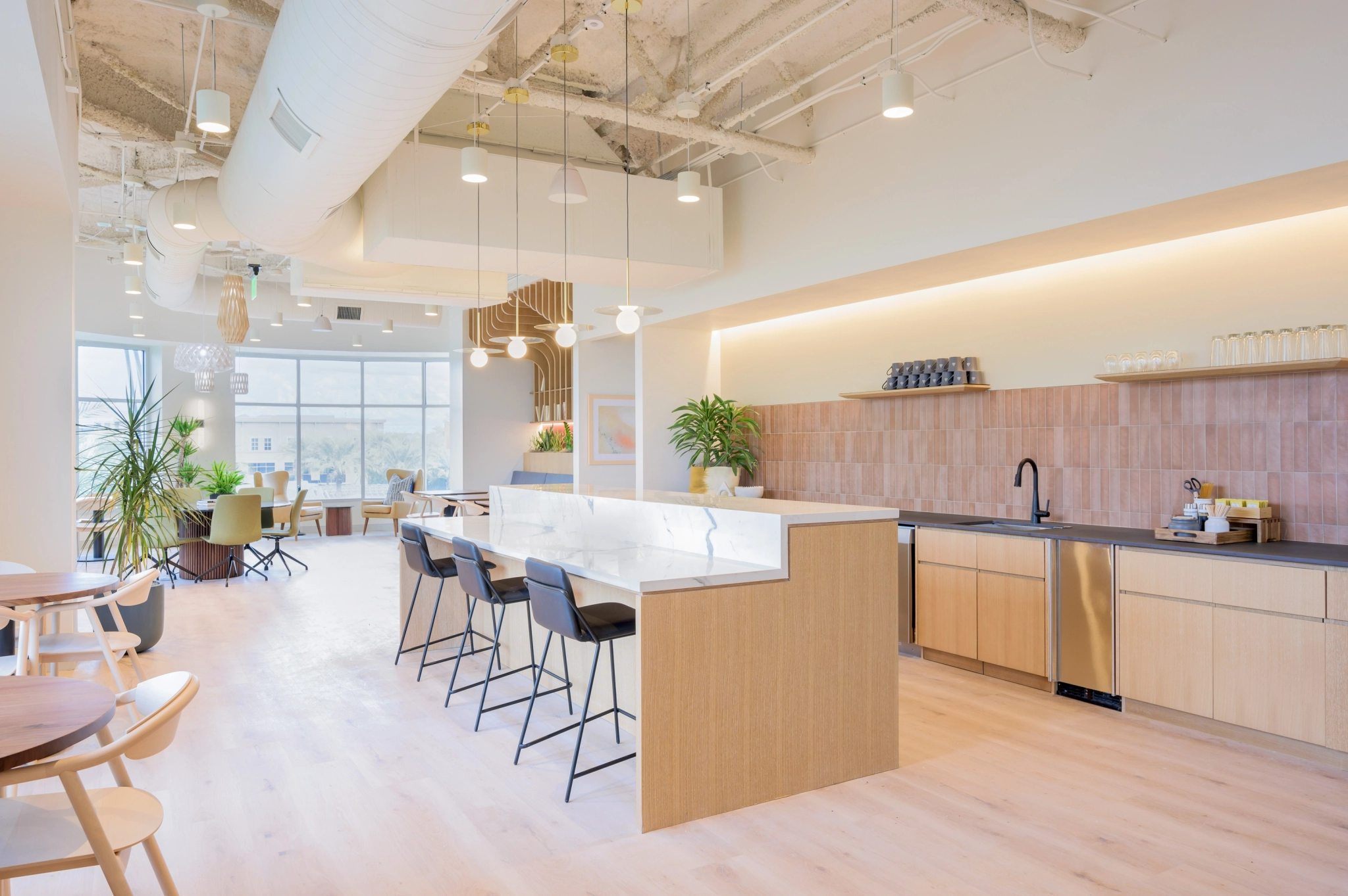 A modern coworking kitchen features a long island, bar stools, wooden cabinetry, and hanging lights. Large windows and plants enhance the bright, open office space.