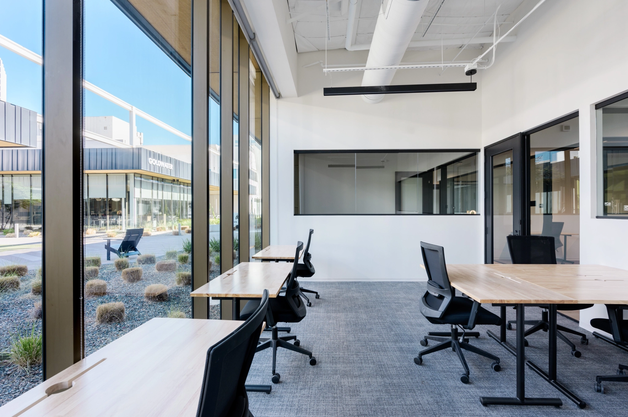 Moderne werkruimte met grote ramen, houten bureaus en ergonomische stoelen. Het kantoor biedt uitzicht naar buiten op een aangelegde ruimte vol planten, waardoor een inspirerende setting ontstaat voor elke vergaderruimte of werkomgeving.